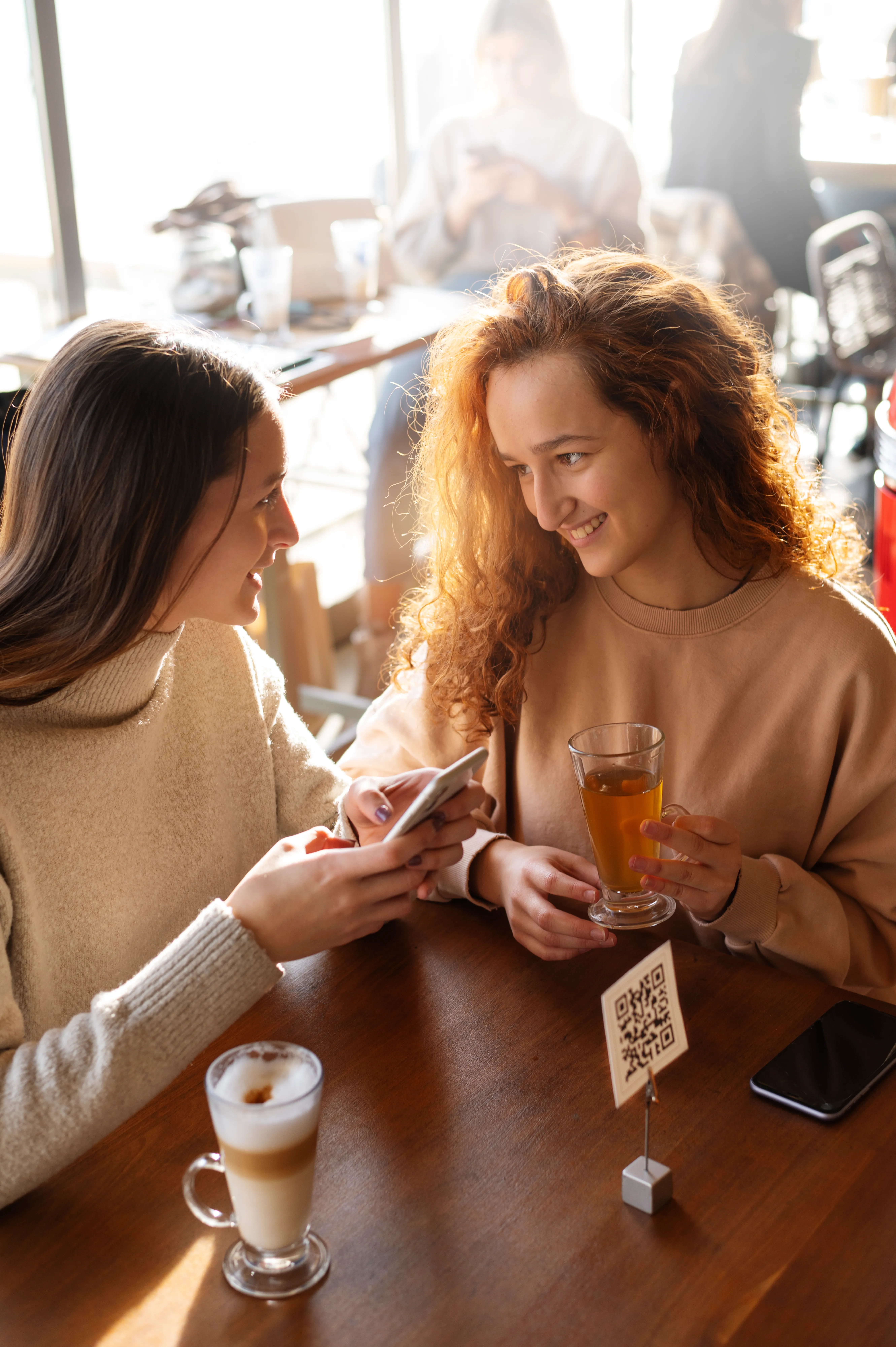  Duas mulheres estão sentadas em uma mesa de café, sorrindo e conversando. A mulher à esquerda segura um celular, enquanto a mulher à direita segura um copo com uma bebida. Na mesa, há um copo de café e um suporte com um cartão que contém um código QR. Ao fundo, outras pessoas estão sentadas e interagindo. A luz natural ilumina o ambiente.