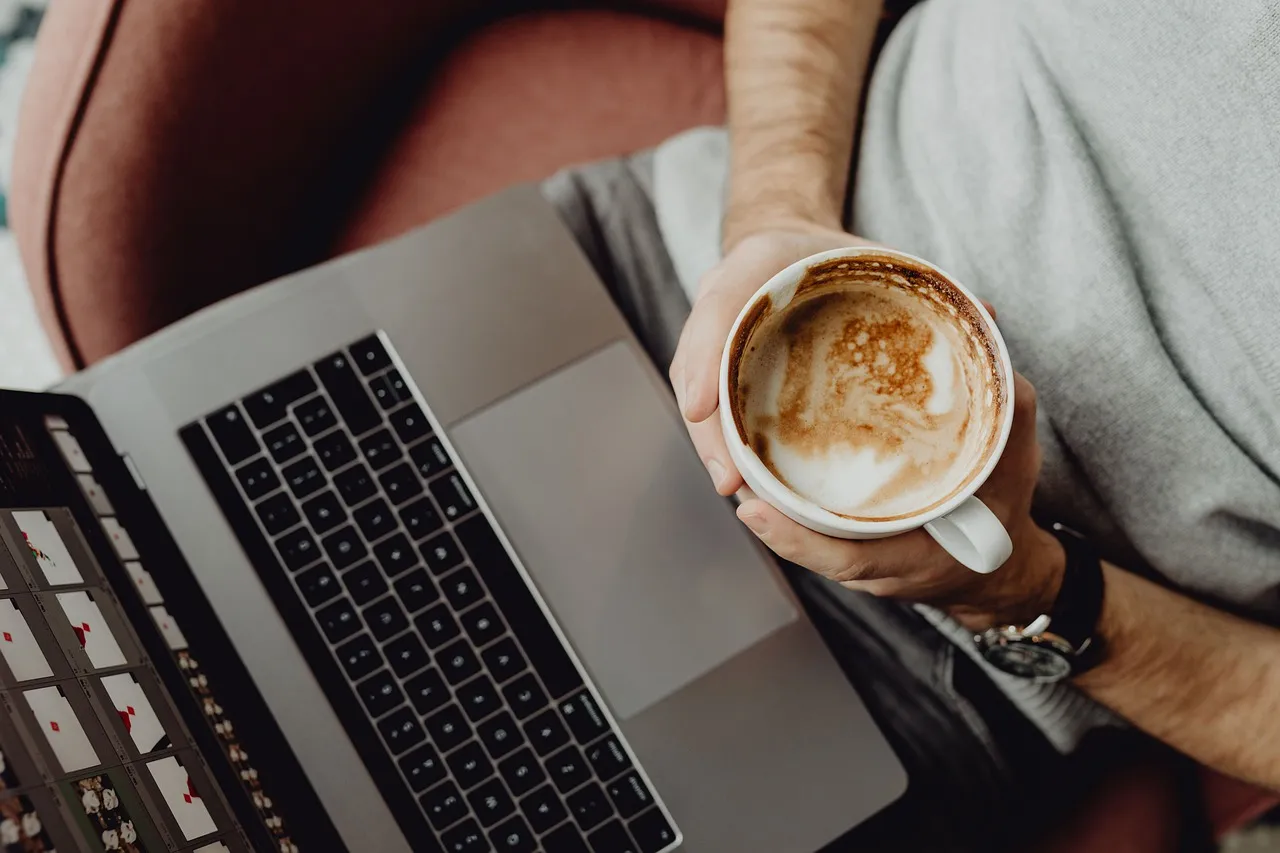 #FotoDescrição: Pessoa segurando uma xícara de café enquanto trabalha em um laptop, simbolizando flexibilidade e trabalho remoto.