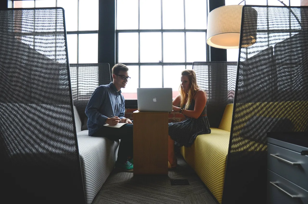 #FotoDescrição: Duas pessoas sentadas em sofás com um laptop aberto em uma mesa, conversando em um ambiente de trabalho moderno.