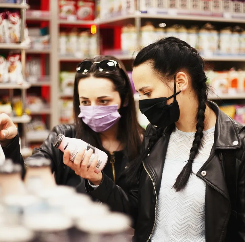 #FotoDescrição: Duas pessoas de máscara examinando um produto em uma prateleira de supermercado, avaliando as informações do rótulo.