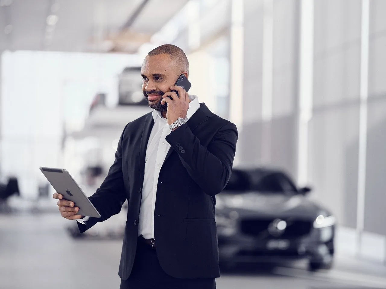 #FotoDescrição: Homem de negócios em um showroom de carros, usando terno preto e conversando ao celular enquanto segura um tablet com a outra mão.