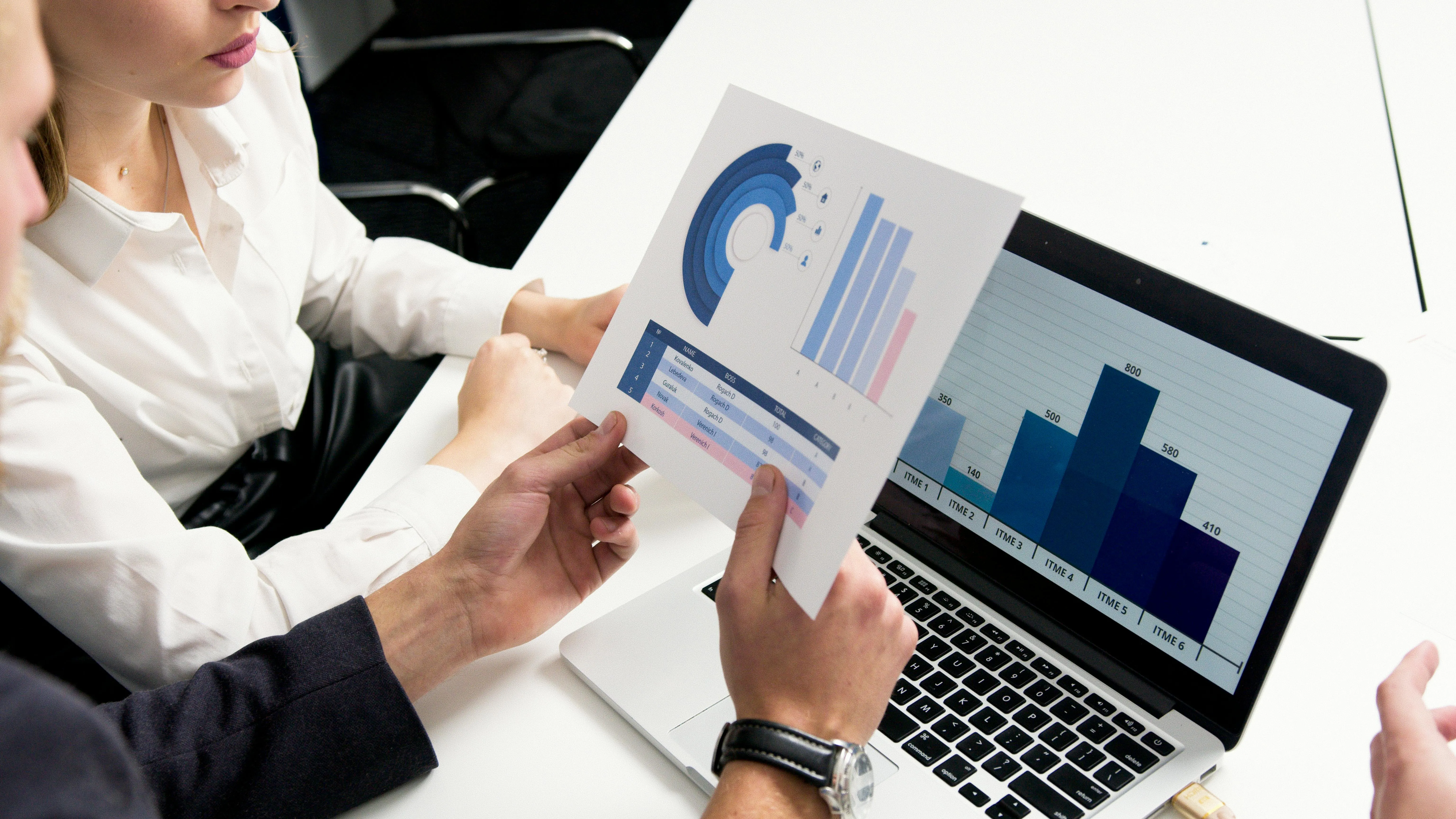#FotoDescrição Duas pessoas em uma mesa de escritório analisando dados financeiros. Uma delas segura uma folha com gráficos circulares e a outra observa um gráfico de barras exibido na tela de um notebook.