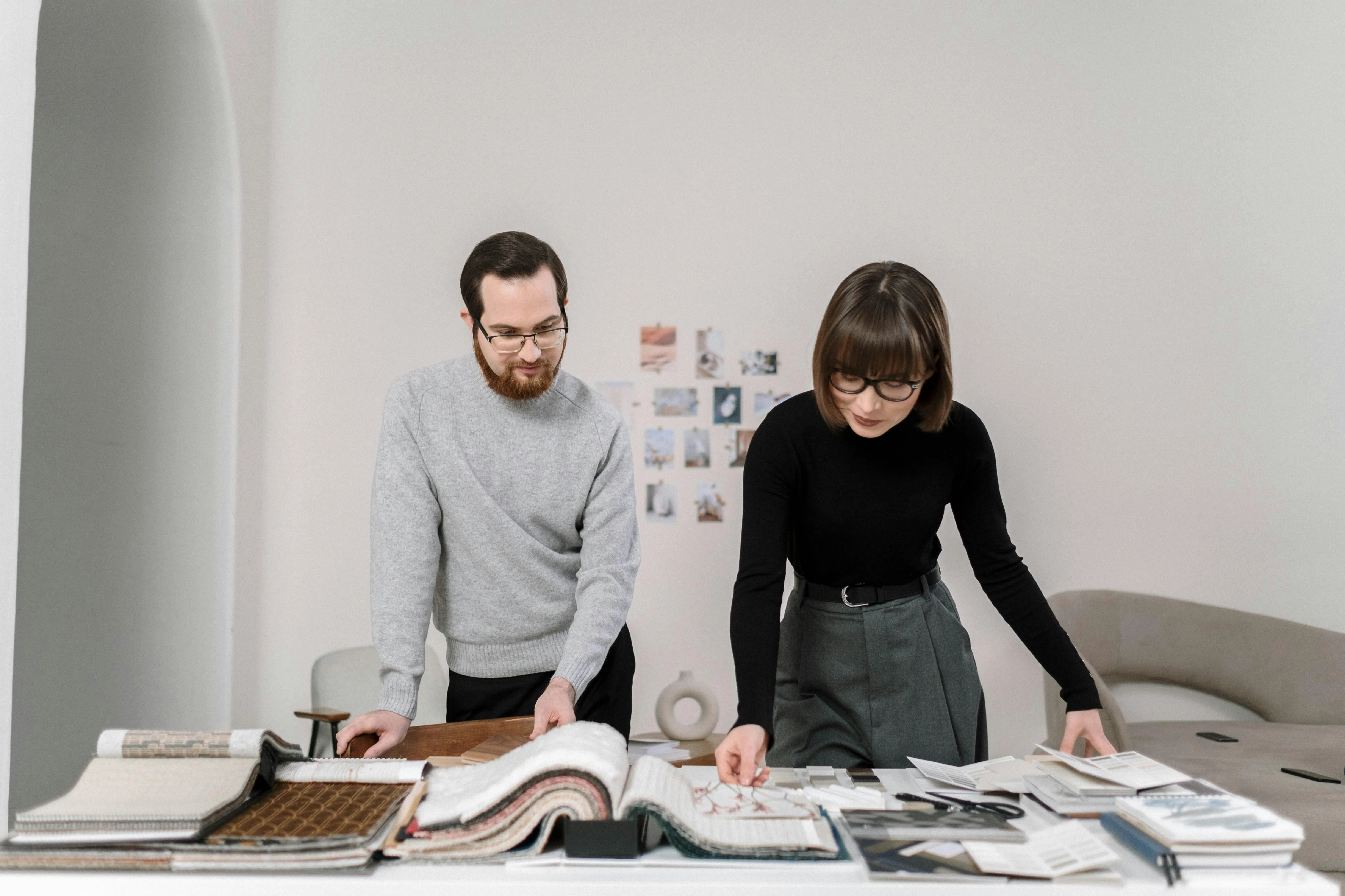 #FotoDescrição Homem e mulher analisam materiais de design ou catálogos sobre uma mesa repleta de amostras, representando uma situação de planejamento visual, criatividade e organização estratégica.
