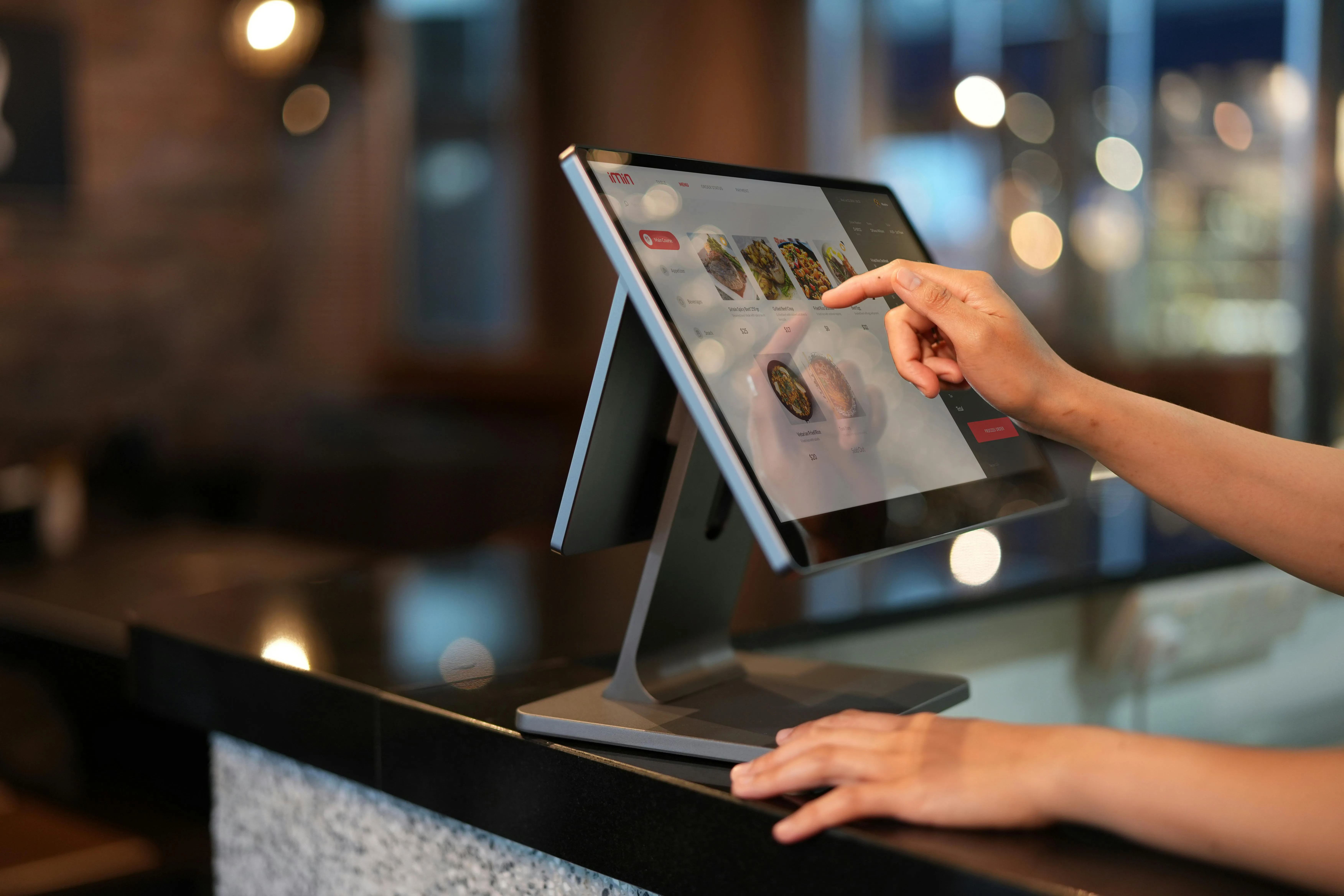 #FotoDescrição Mão tocando a tela de um monitor touchscreen em um balcão moderno, com imagens de pratos exibidas na interface digital.
