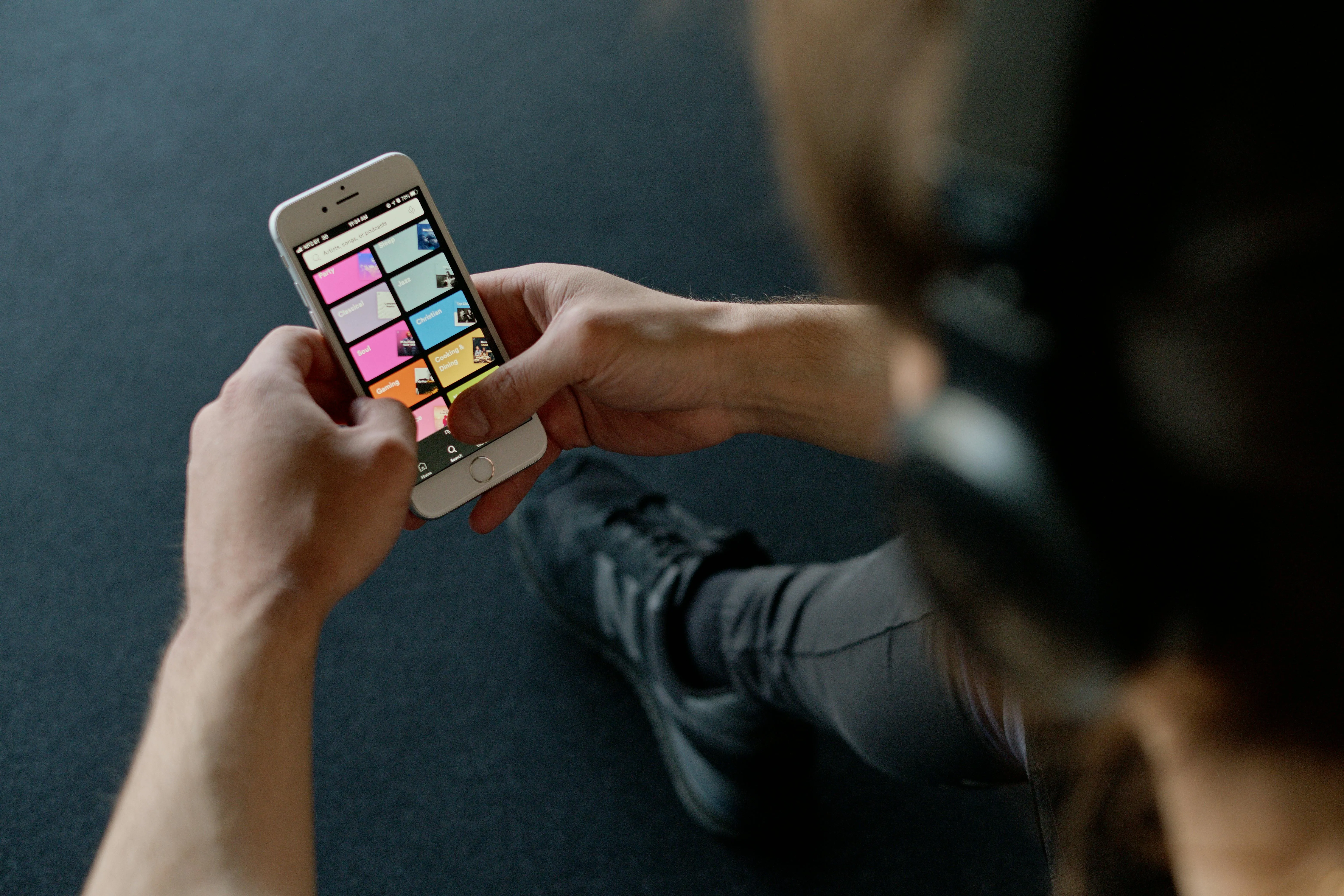 #FotoDescrição: Pessoa segurando um smartphone com fones de ouvido no pescoço, visualizando o aplicativo de música com categorias coloridas na tela.