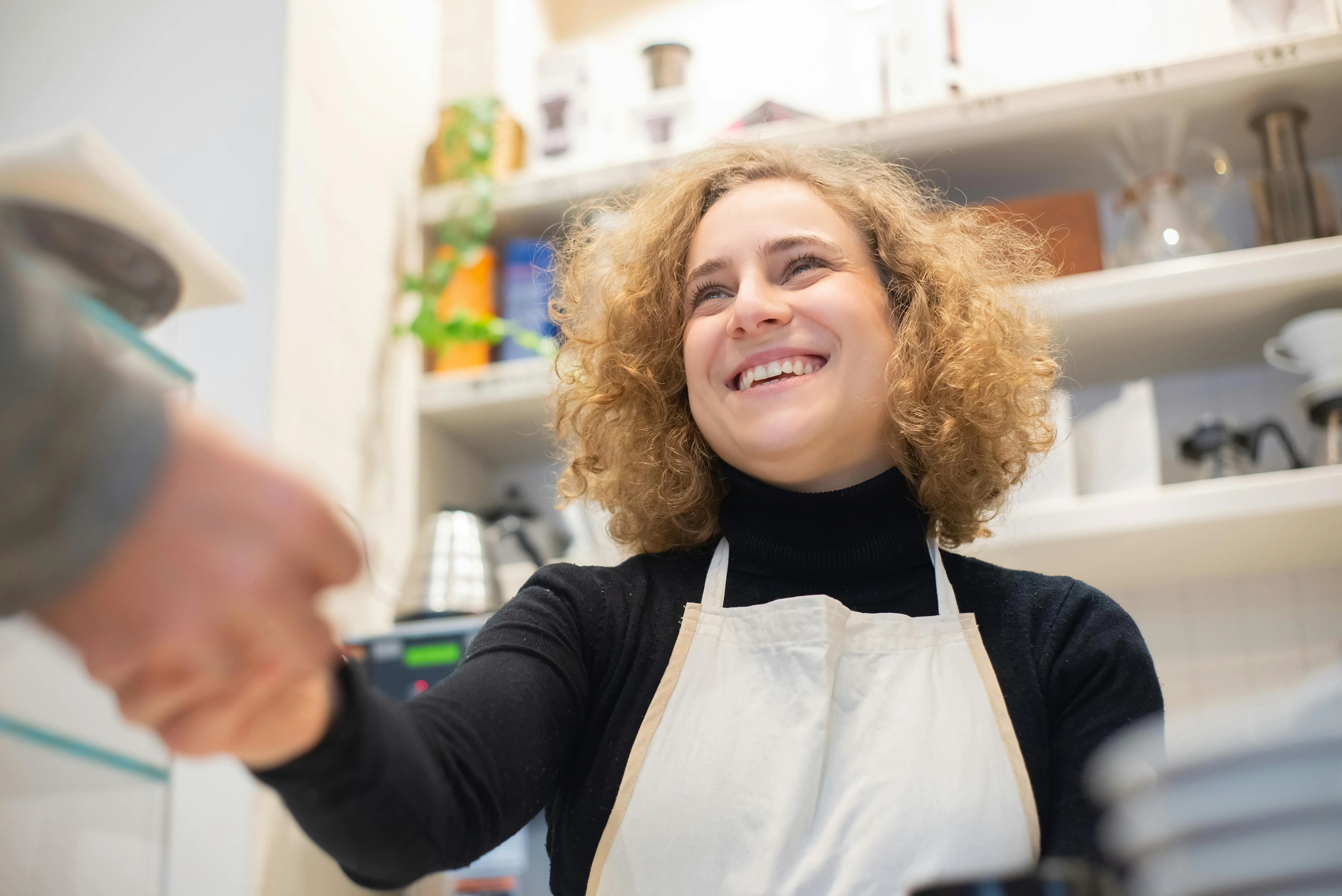 #FotoDescrição Funcionária de um café sorrindo enquanto aperta a mão de um cliente, usando avental claro sobre roupa preta.