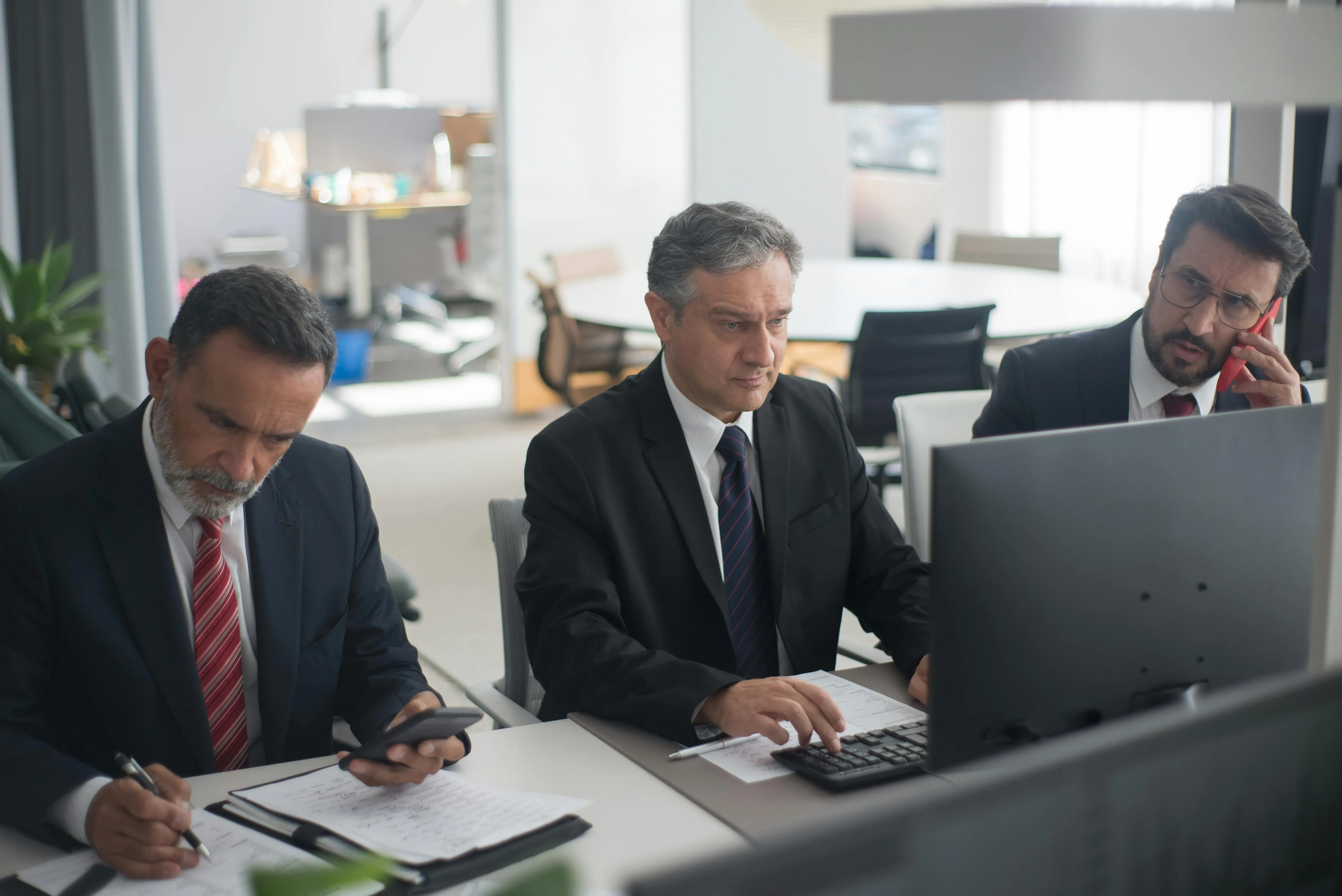 #FotoDescrição: Três homens de terno sentados em um escritório moderno; dois preenchem anotações enquanto o terceiro fala ao telefone e observa um monitor.