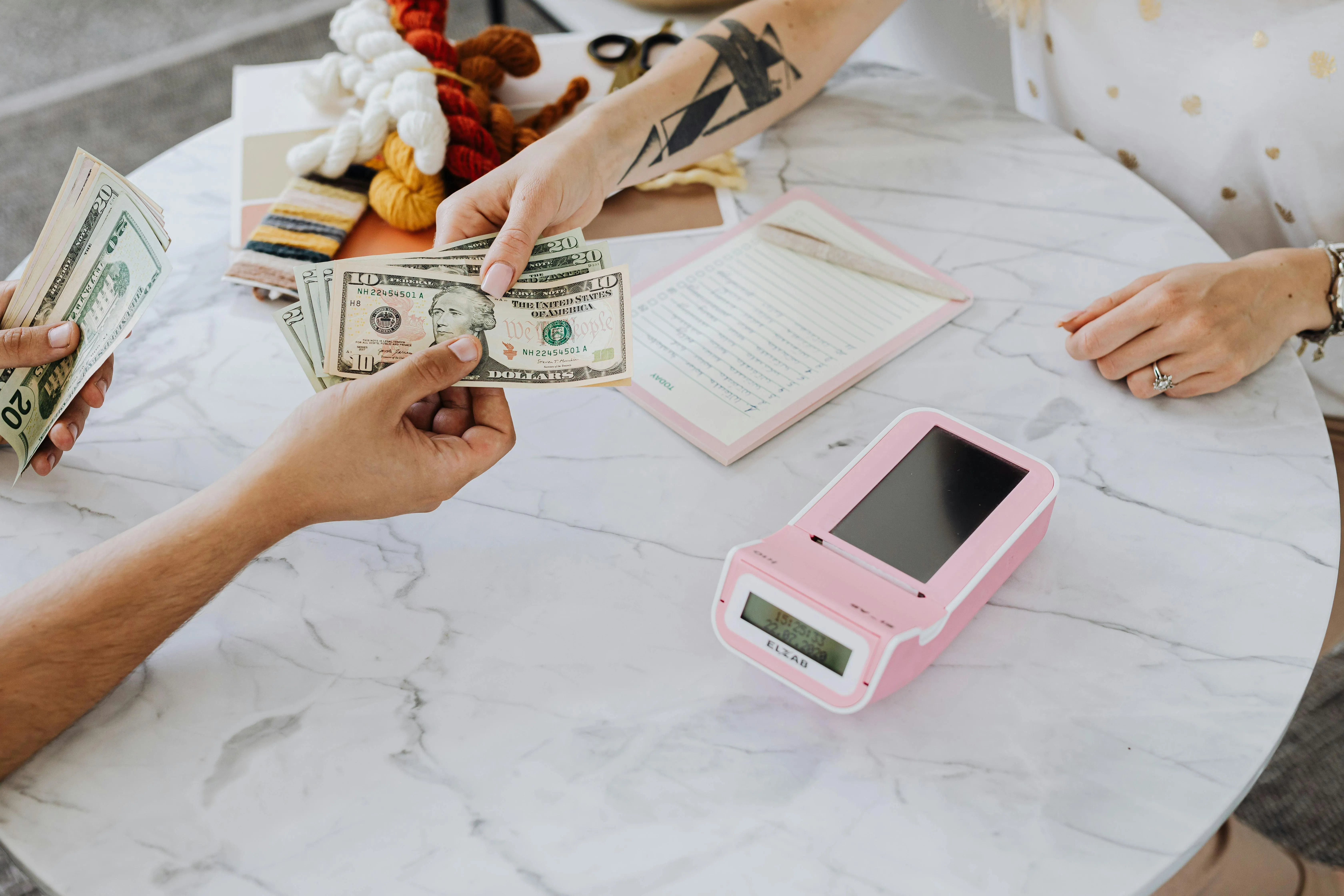 Pessoa entregando notas de dólar em uma mesa com caderno e máquina rosa.