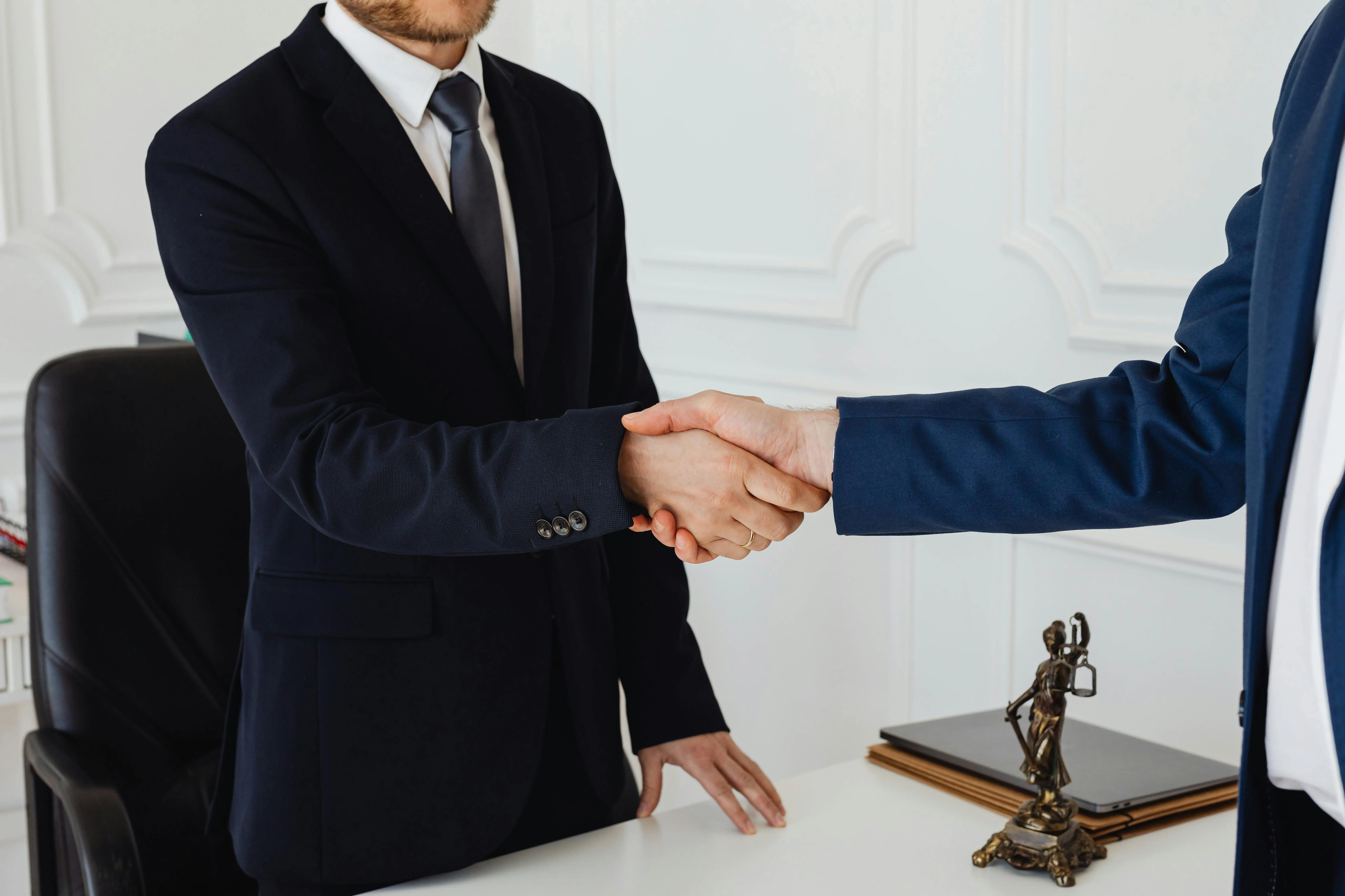 #FotoDescrição: Dois profissionais de terno apertando as mãos em um escritório, simbolizando acordo, confiança e alinhamento empresarial.