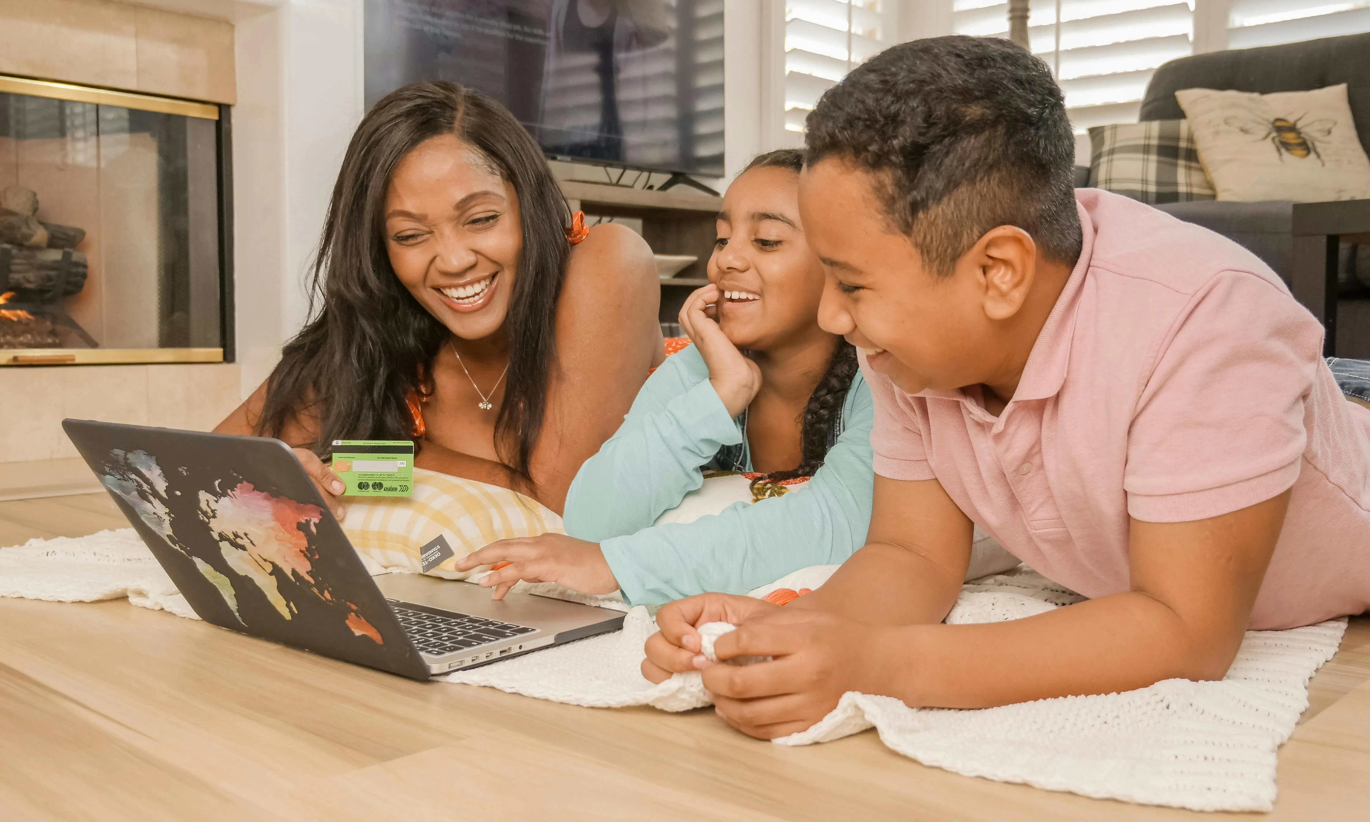 #FotoDescrição: Mãe e duas crianças sorrindo enquanto olham para a tela de um laptop em casa.