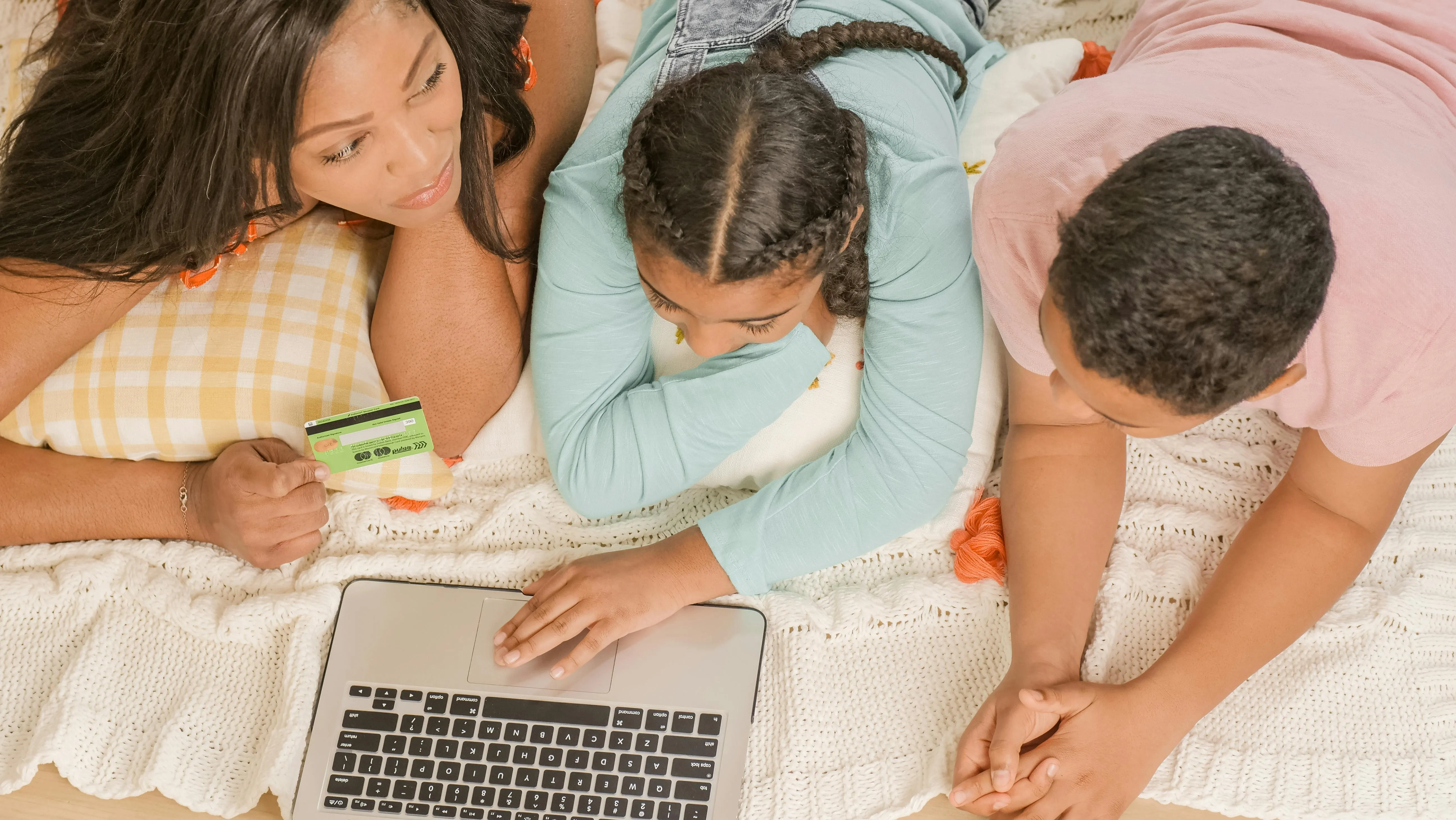 #FotoDescrição Família deitada em um tapete com notebook e cartão de crédito na mão.