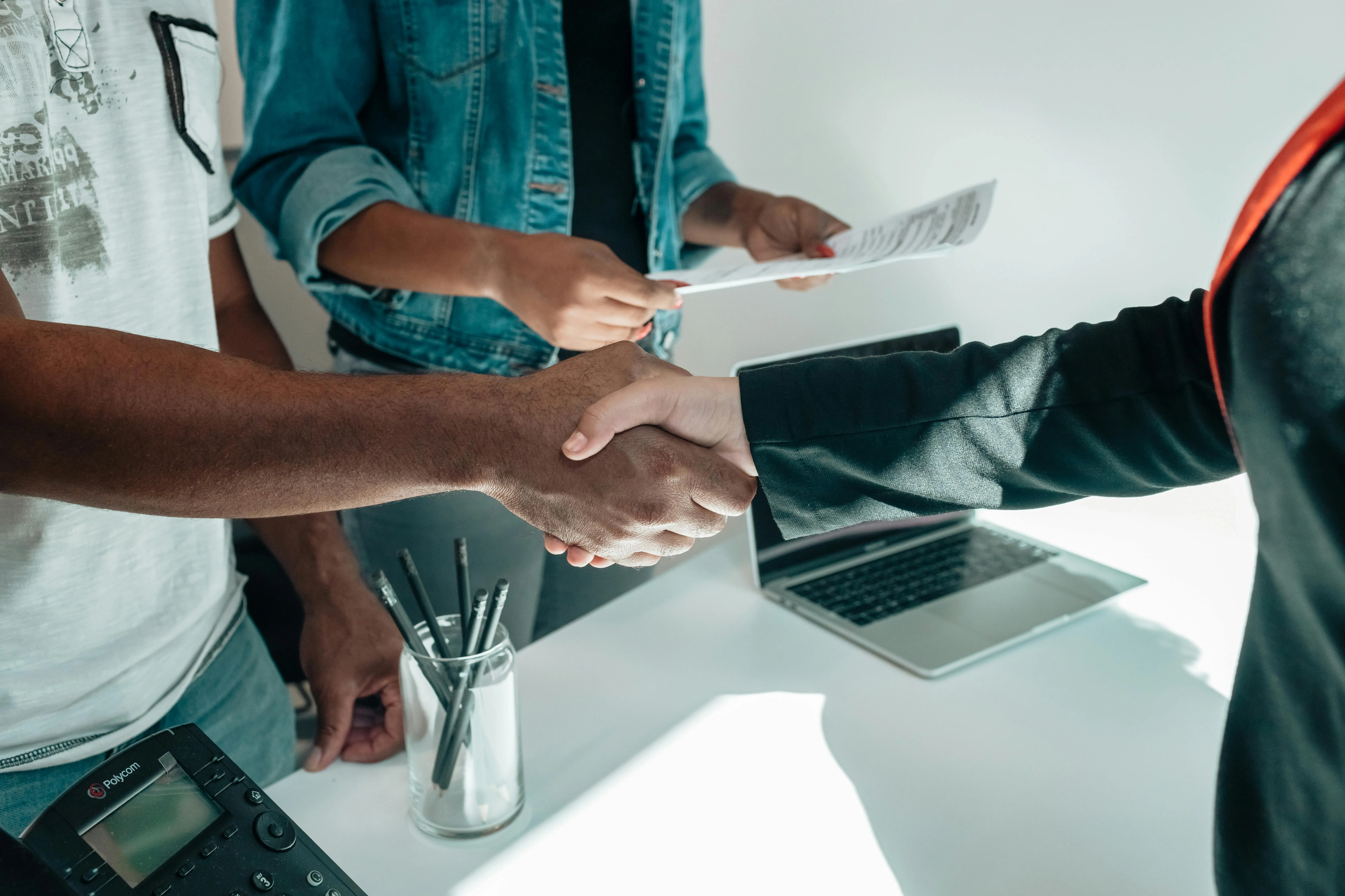 Duas pessoas apertando mãos em ambiente de negócios, com mesa e laptop ao fundo.