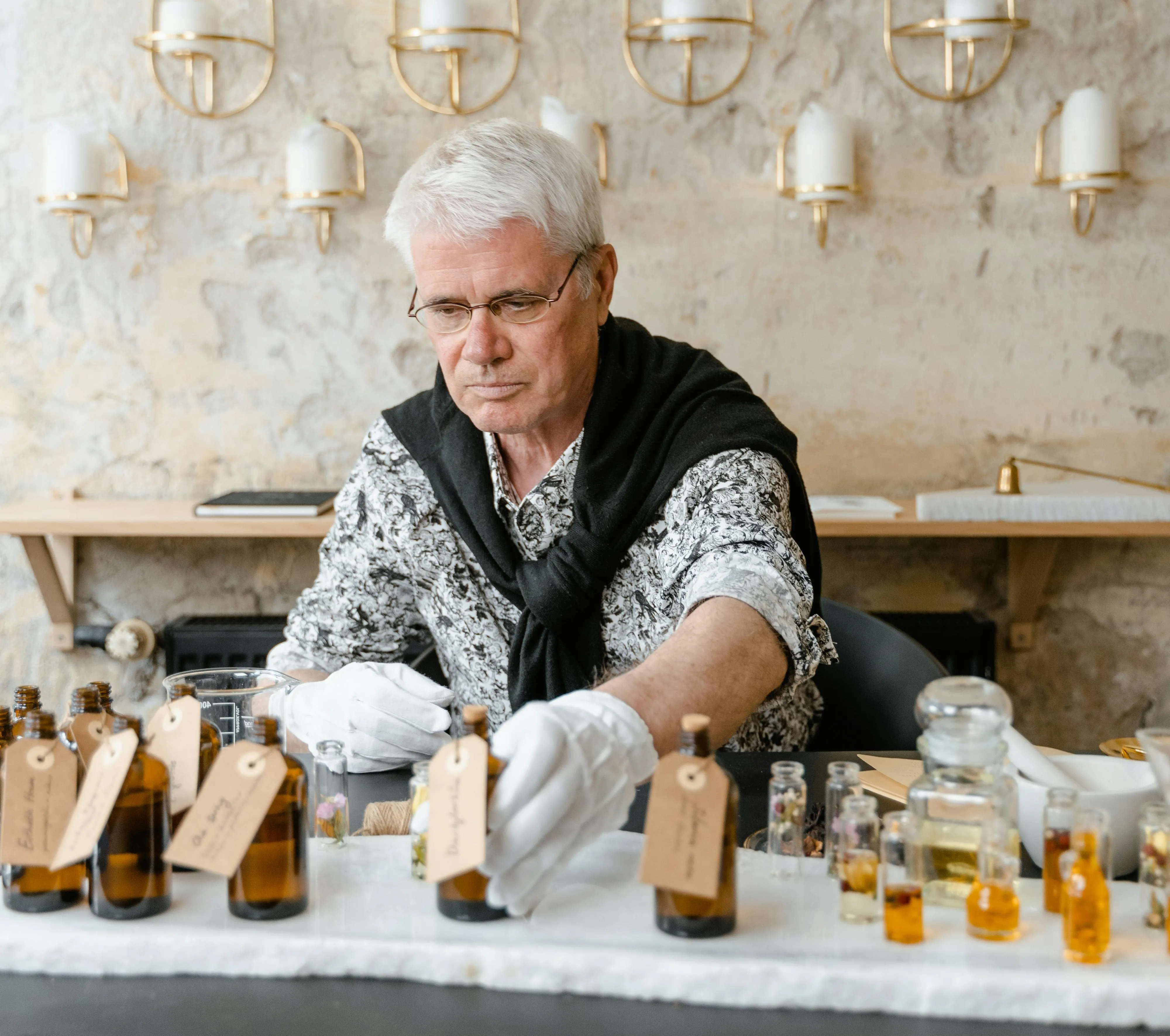 #FotoDescrição: Um homem idoso manuseando frascos com líquidos em um ambiente iluminado, representando a fabricação artesanal ou análise química.