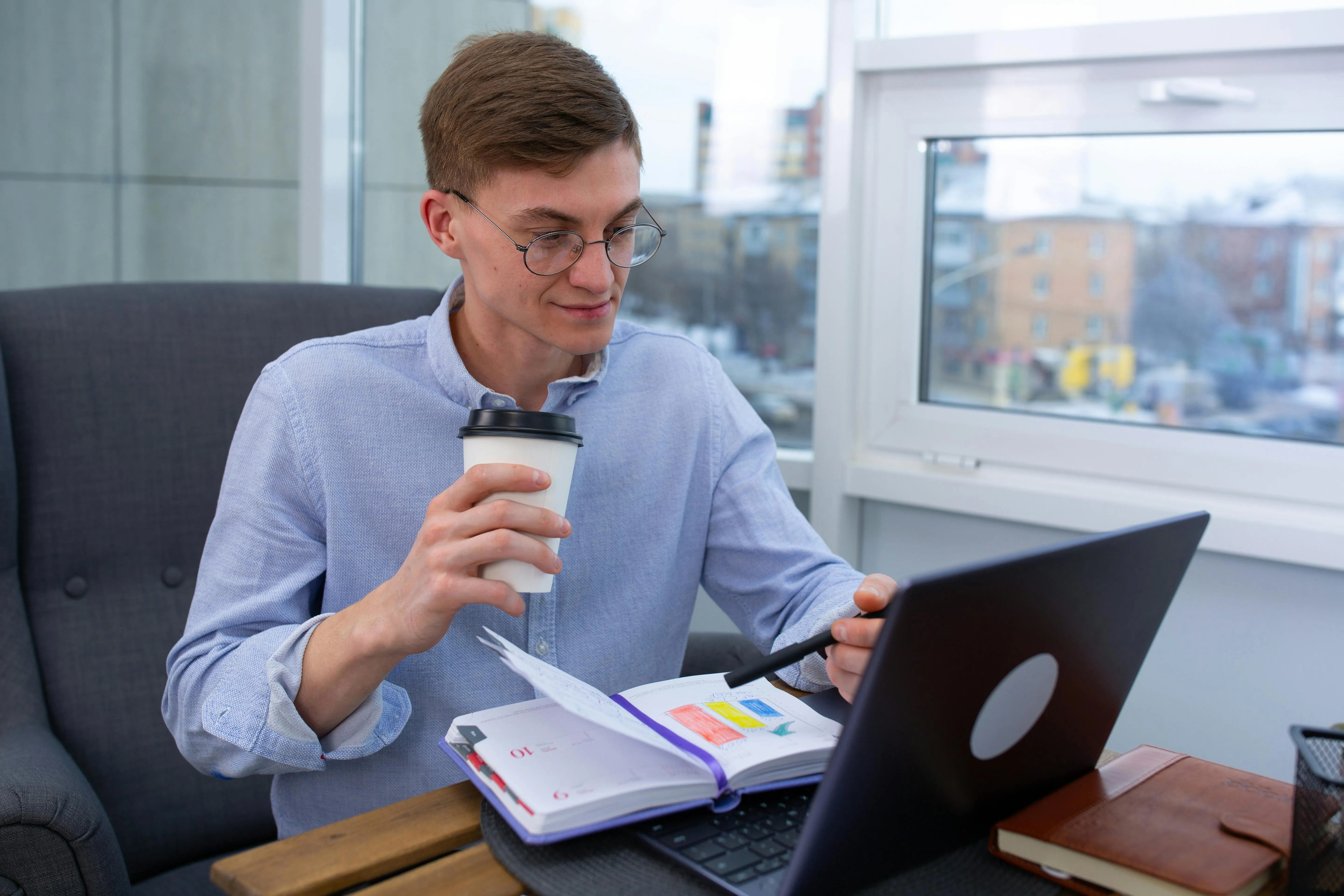 Jovem trabalha no notebook com café e agenda aberta.