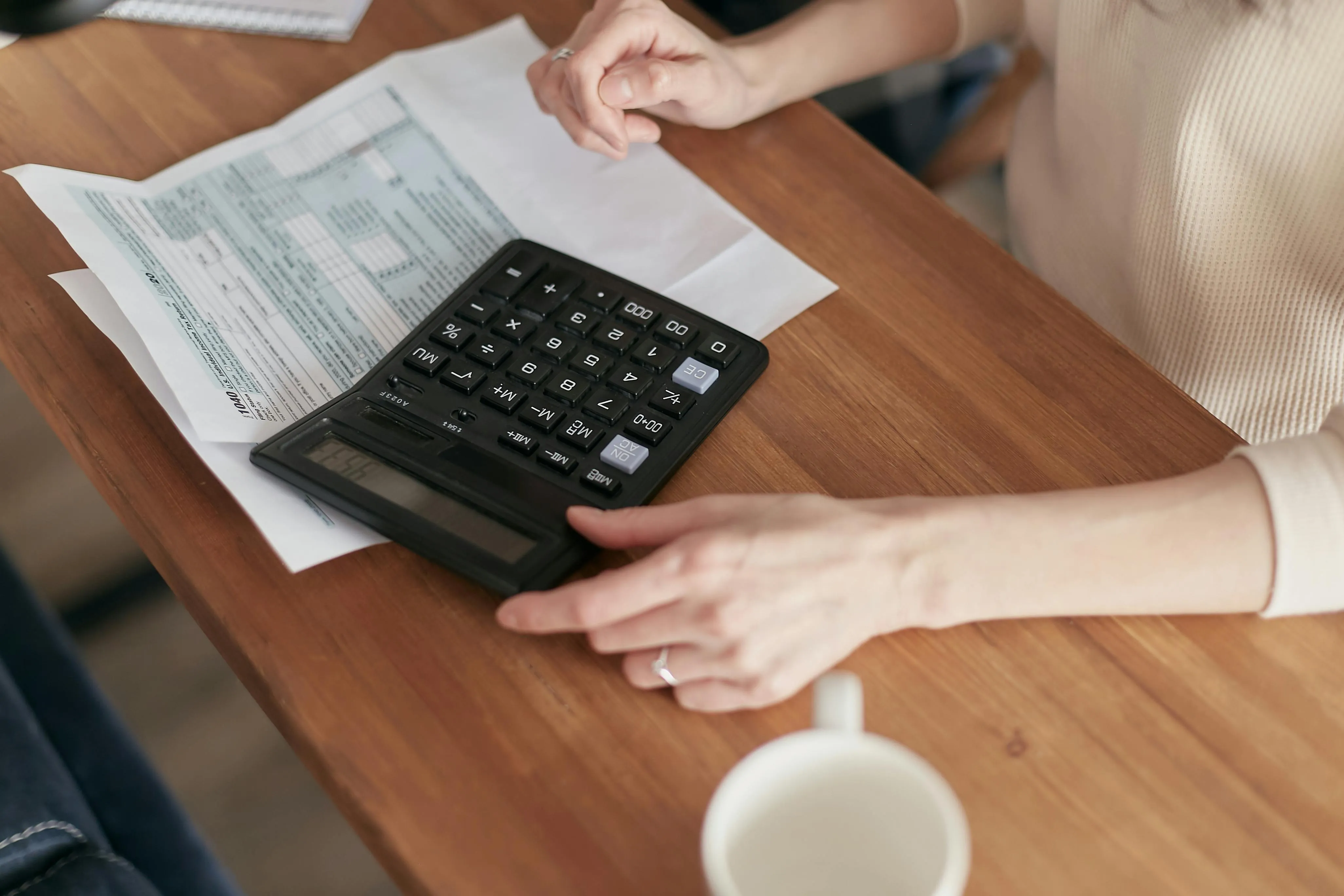 #FotoDescrição: Pessoa usando uma calculadora sobre documentos e boletos em uma mesa de madeira, ao lado de uma xícara de café.