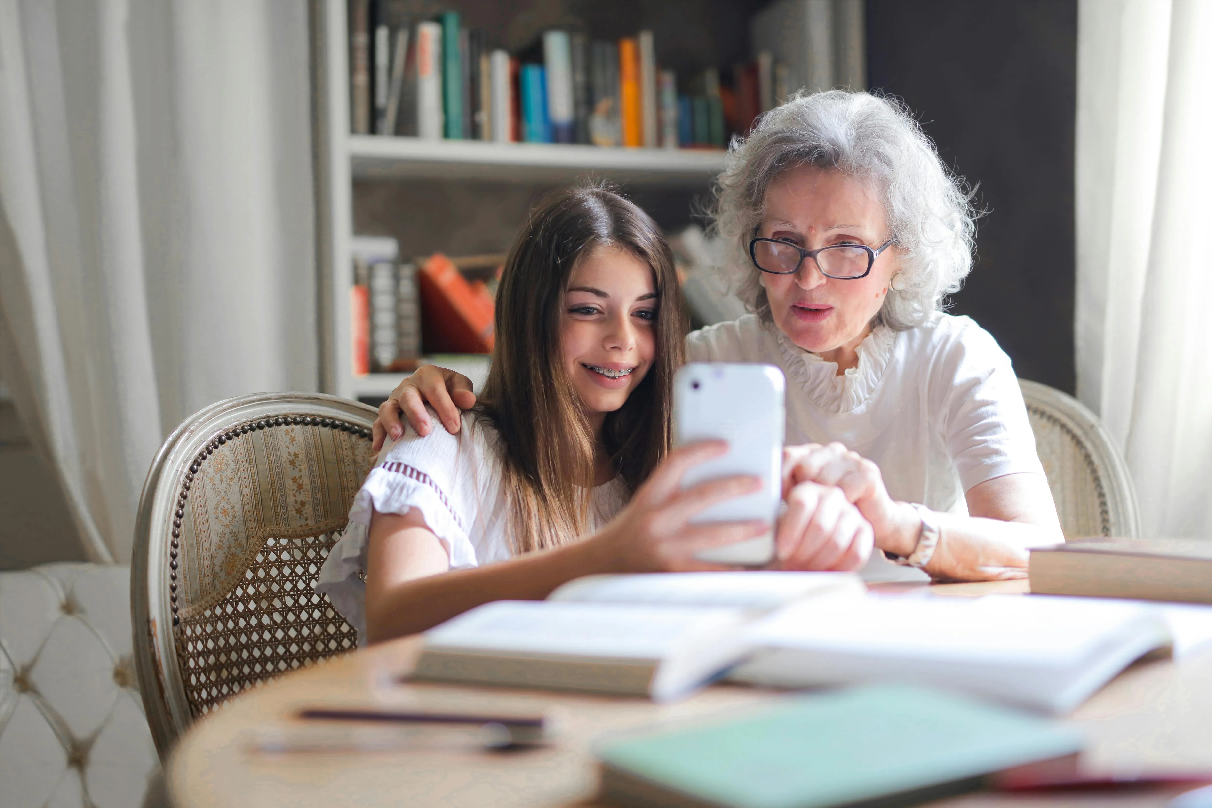 #FotoDescrição Idosa e jovem sentadas juntas, olhando para um celular e sorrindo, com livros ao redor.