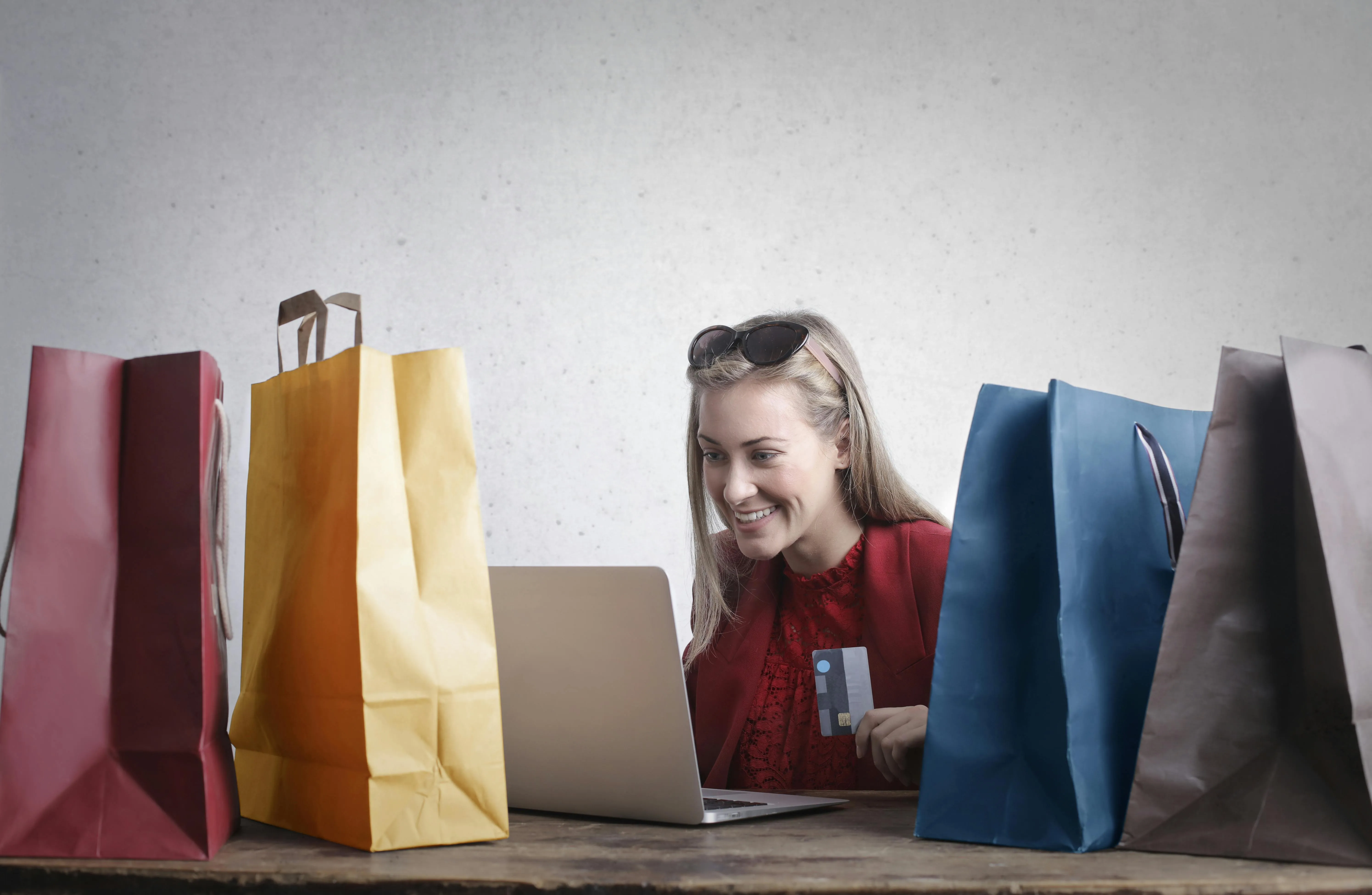 #FotoDescrição: Mulher sorrindo e segurando um cartão de crédito enquanto faz compras online, cercada por sacolas de compras.