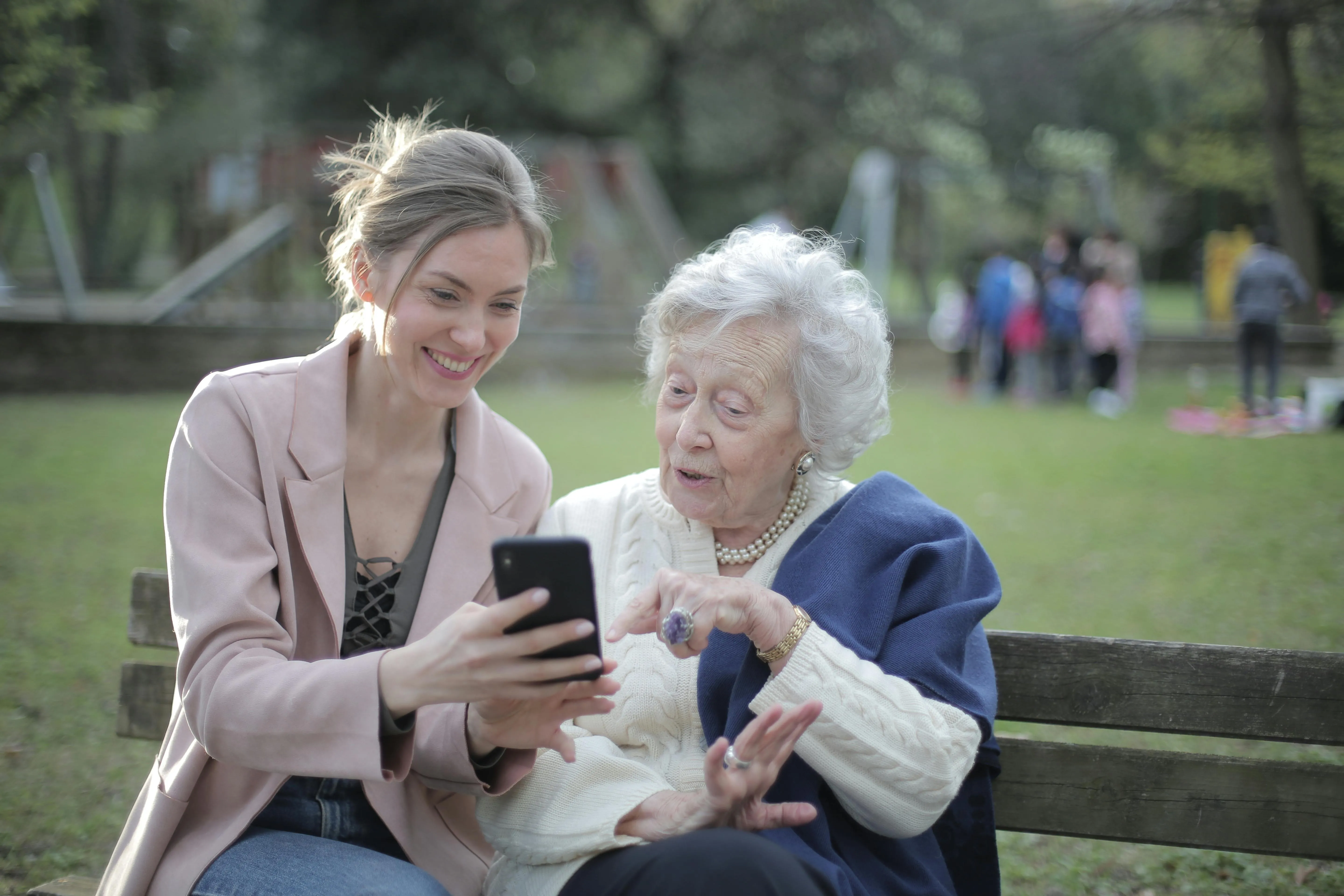 #FotoDescrição: Jovem e idosa sentadas em um banco de praça olhando sorridentes para a tela de um celular.