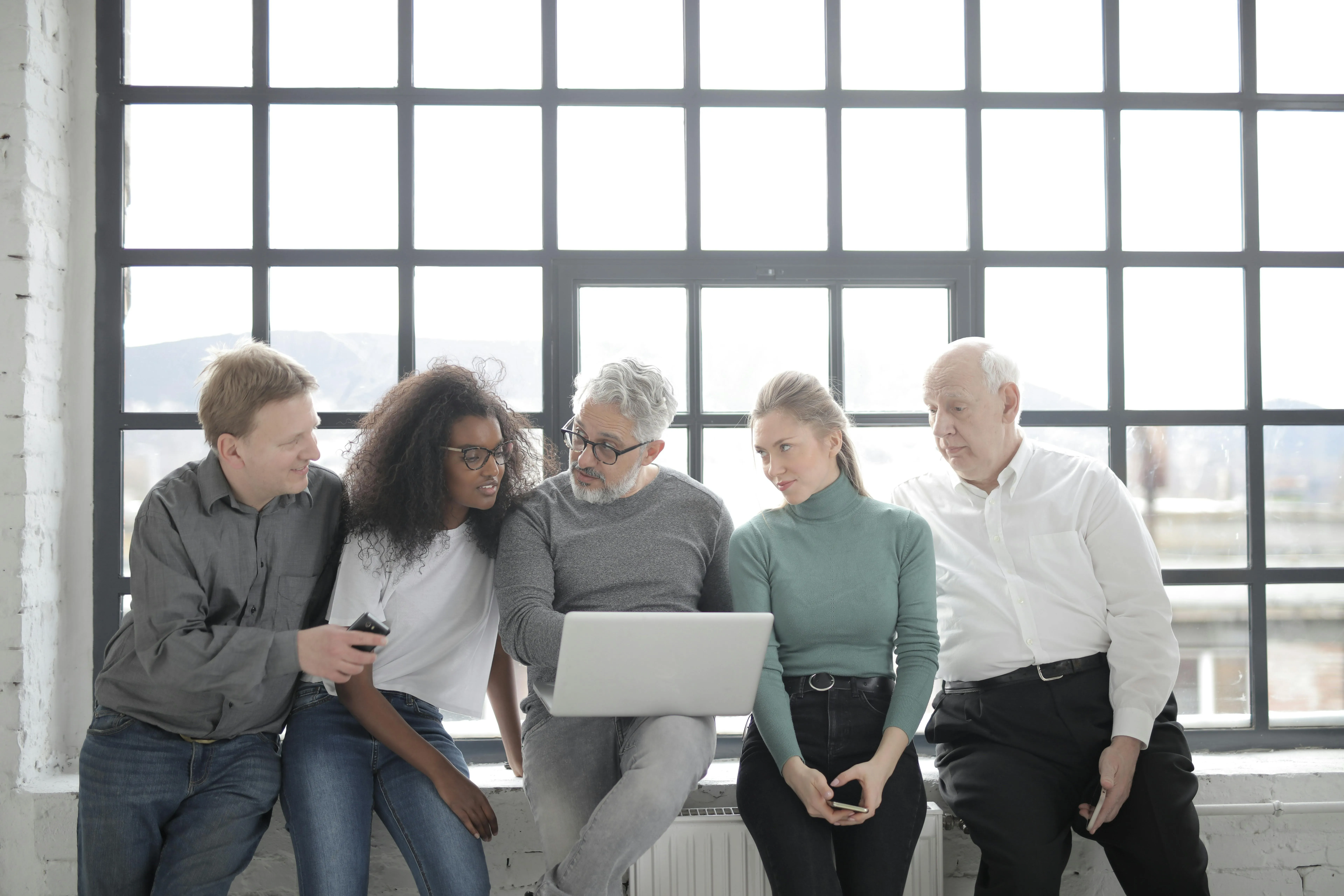 #FotoDescrição: Cinco pessoas de diferentes idades e estilos sentadas lado a lado, colaborando em frente a um notebook com expressão de foco e interesse.