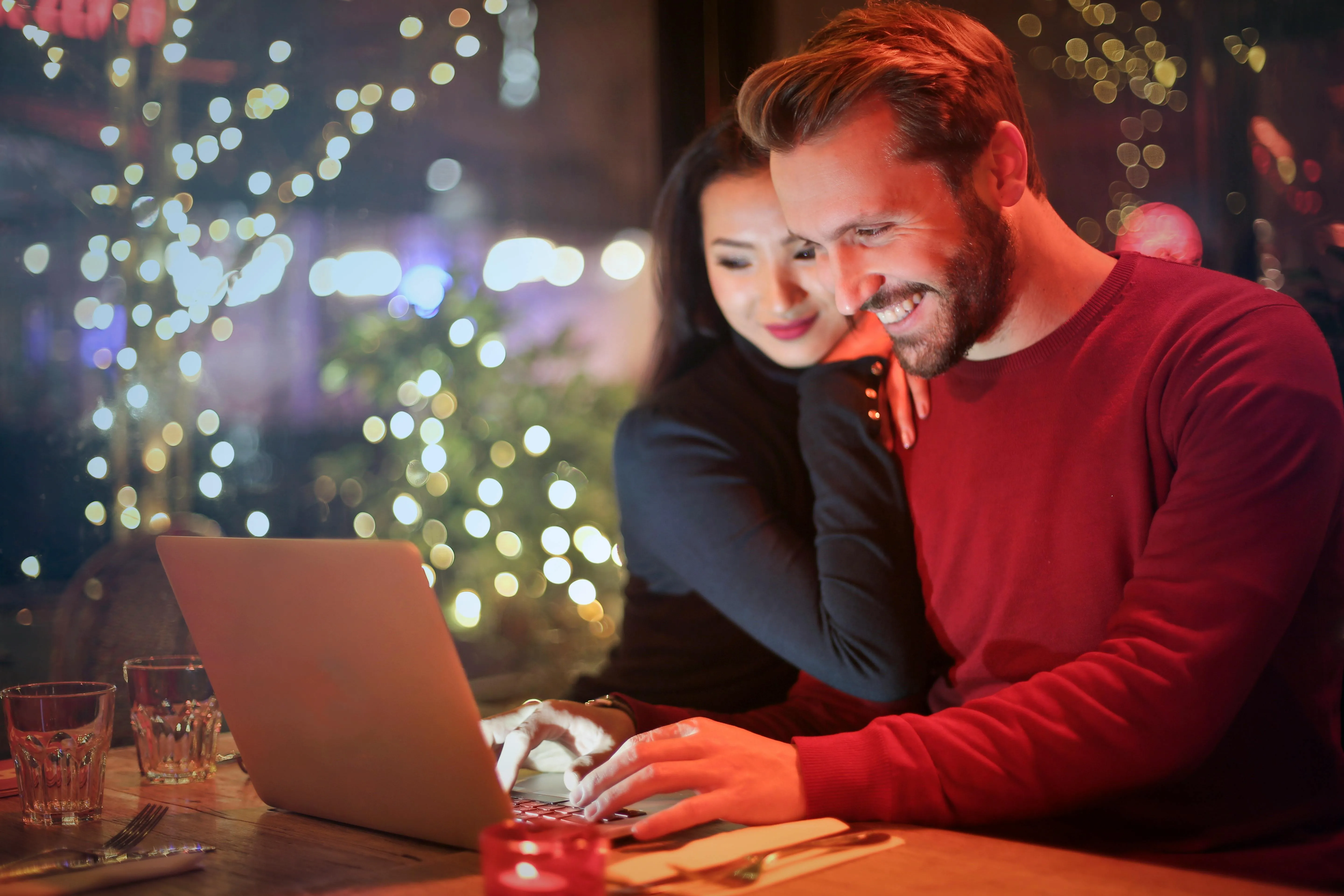 #FotoDescrição: Casal sentado em um ambiente iluminado por luzes de decoração, sorrindo enquanto navegam juntos em um laptop.