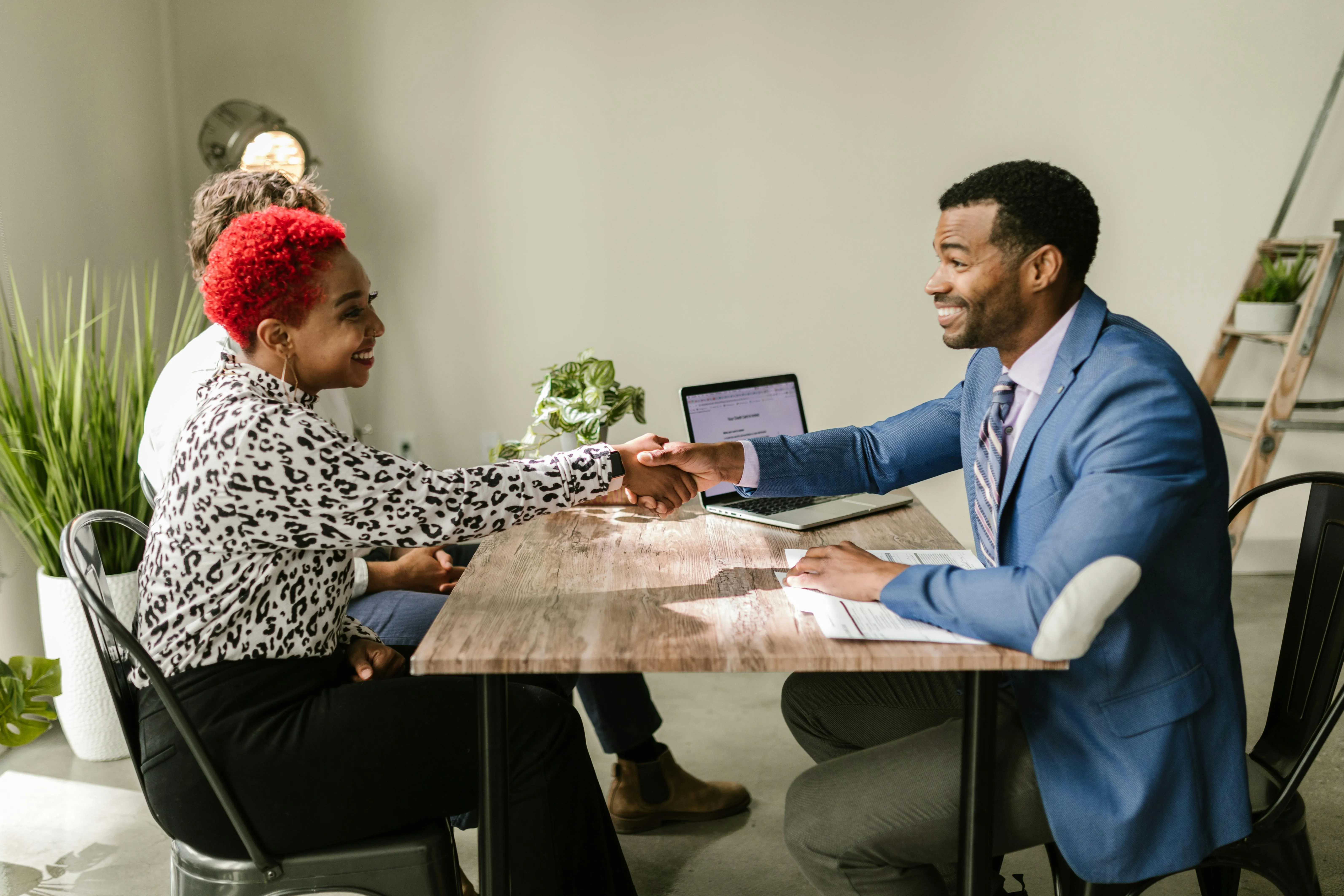 #FotoDescrição: Duas pessoas sorrindo e apertando as mãos em uma mesa de reunião, com computador e plantas ao fundo.