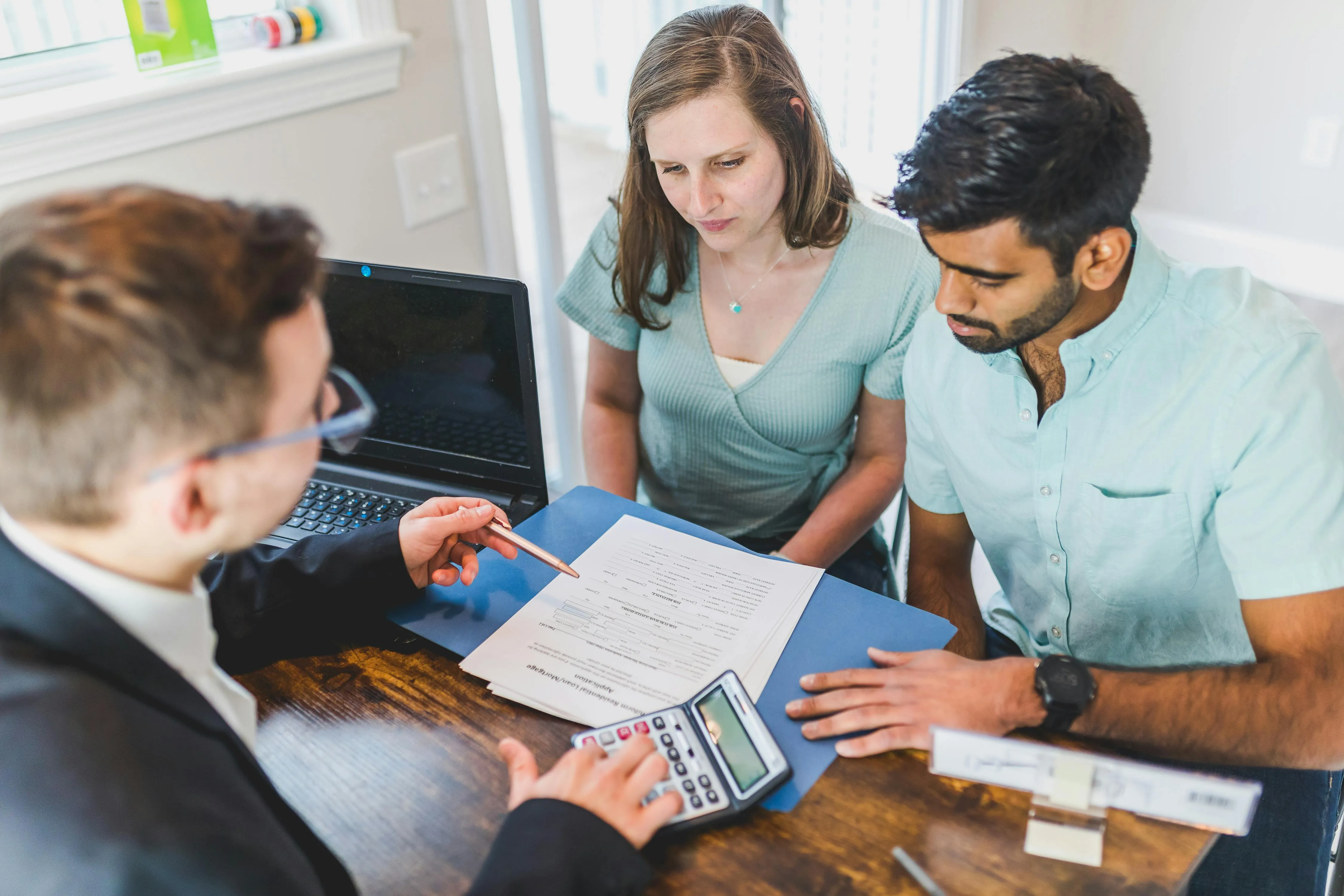 #FotoDescrição: Três pessoas reunidas em uma mesa, discutindo documentos com uma calculadora e um laptop ao fundo.