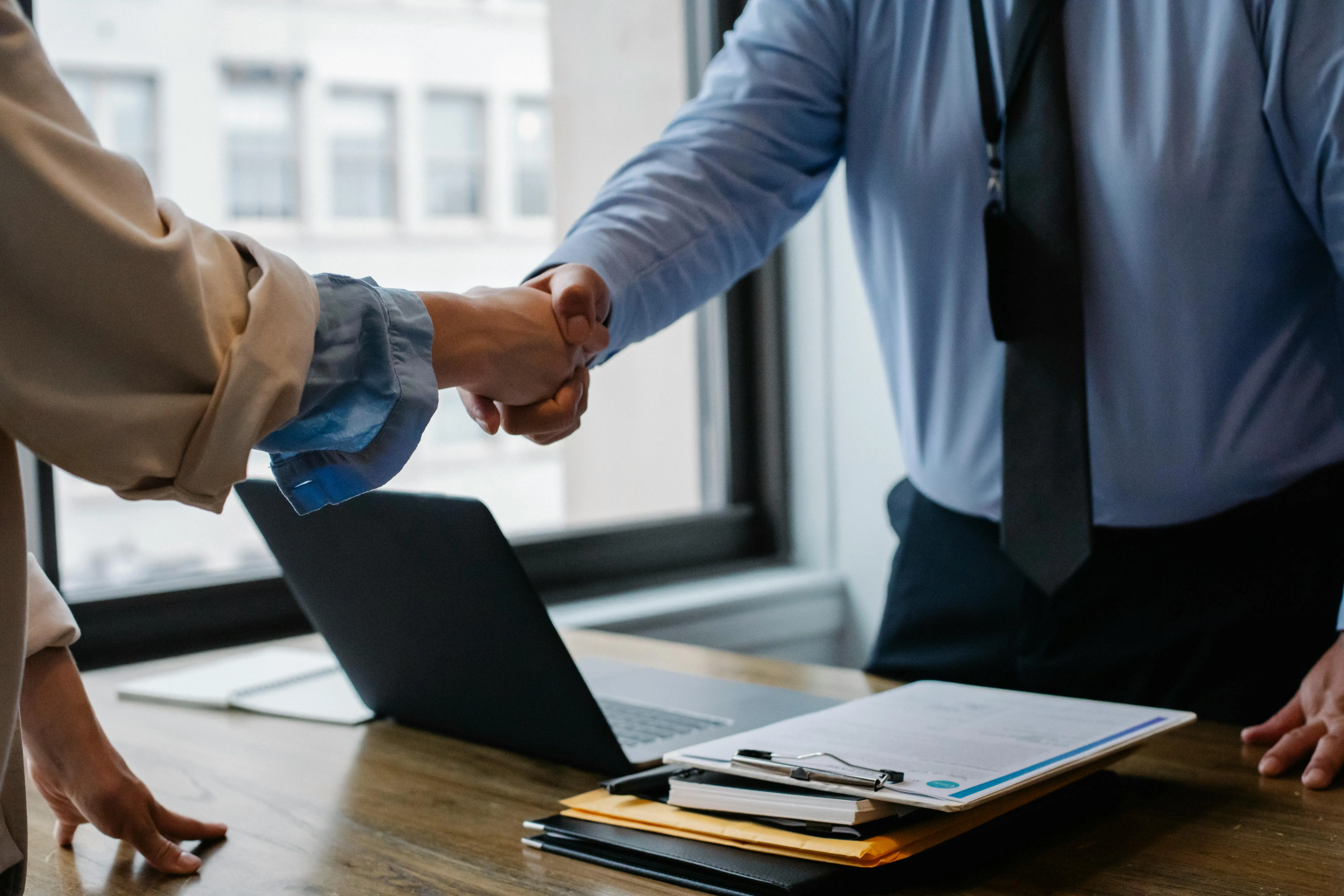 #FotoDescrição Duas pessoas apertando as mãos sobre uma mesa de escritório com laptop e documentos, simbolizando fechamento de parceria ou venda de produto digital em ambiente corporativo.