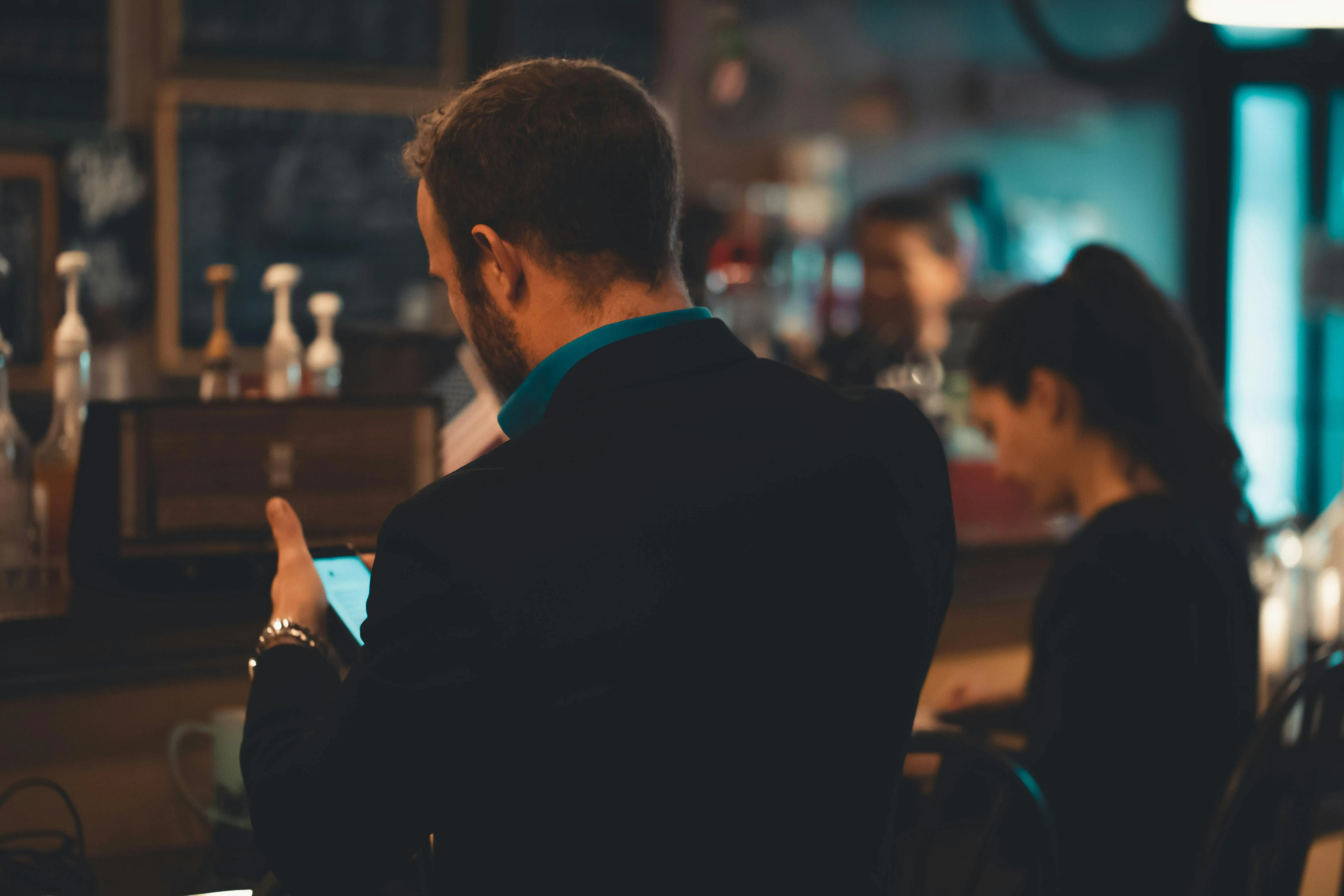 Homem de terno azul em bar, olhando para o celular, com pessoas ao fundo.