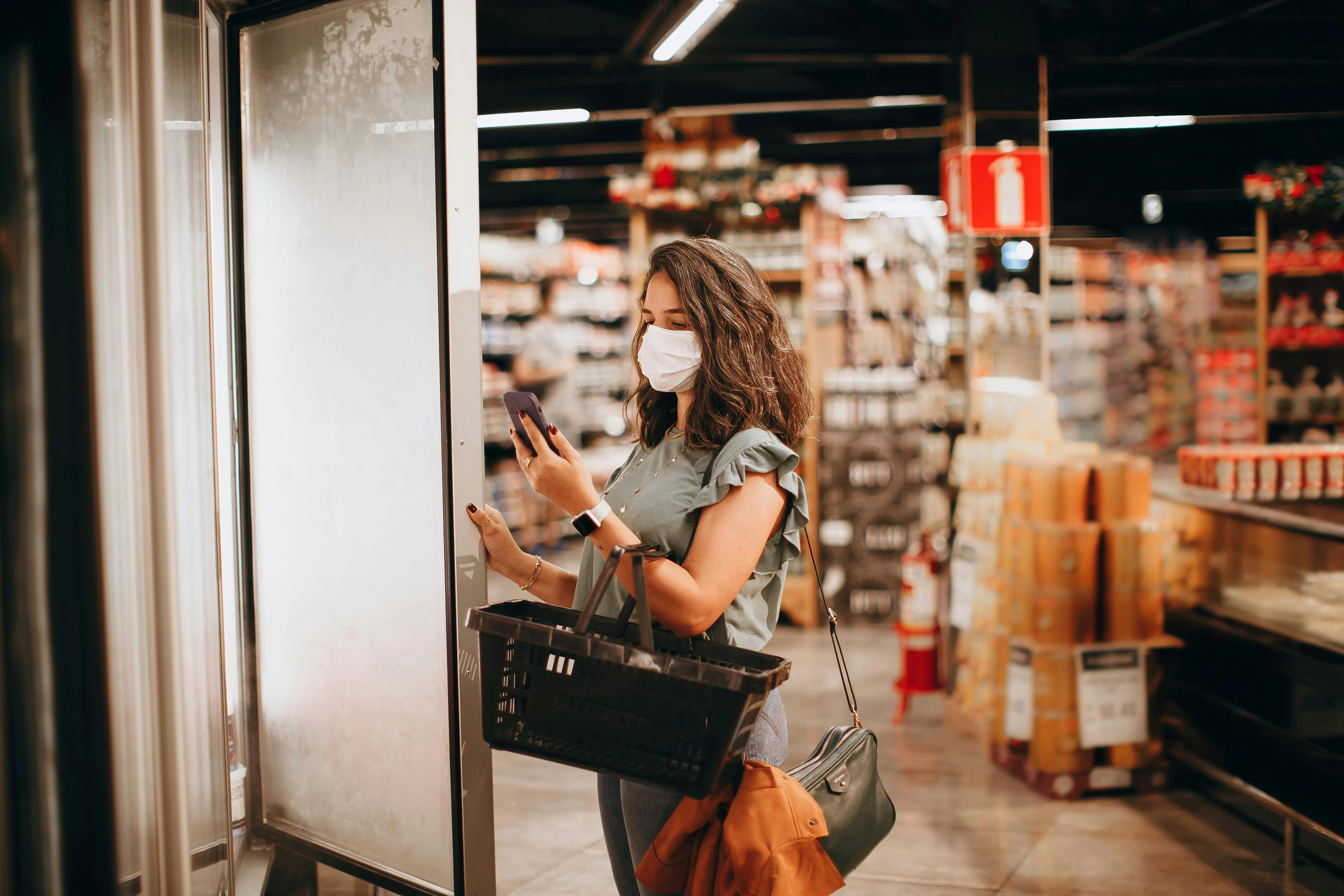 #FotoDescrição: Mulher usando máscara, segurando uma cesta de compras e abrindo uma porta de refrigerador em supermercado.