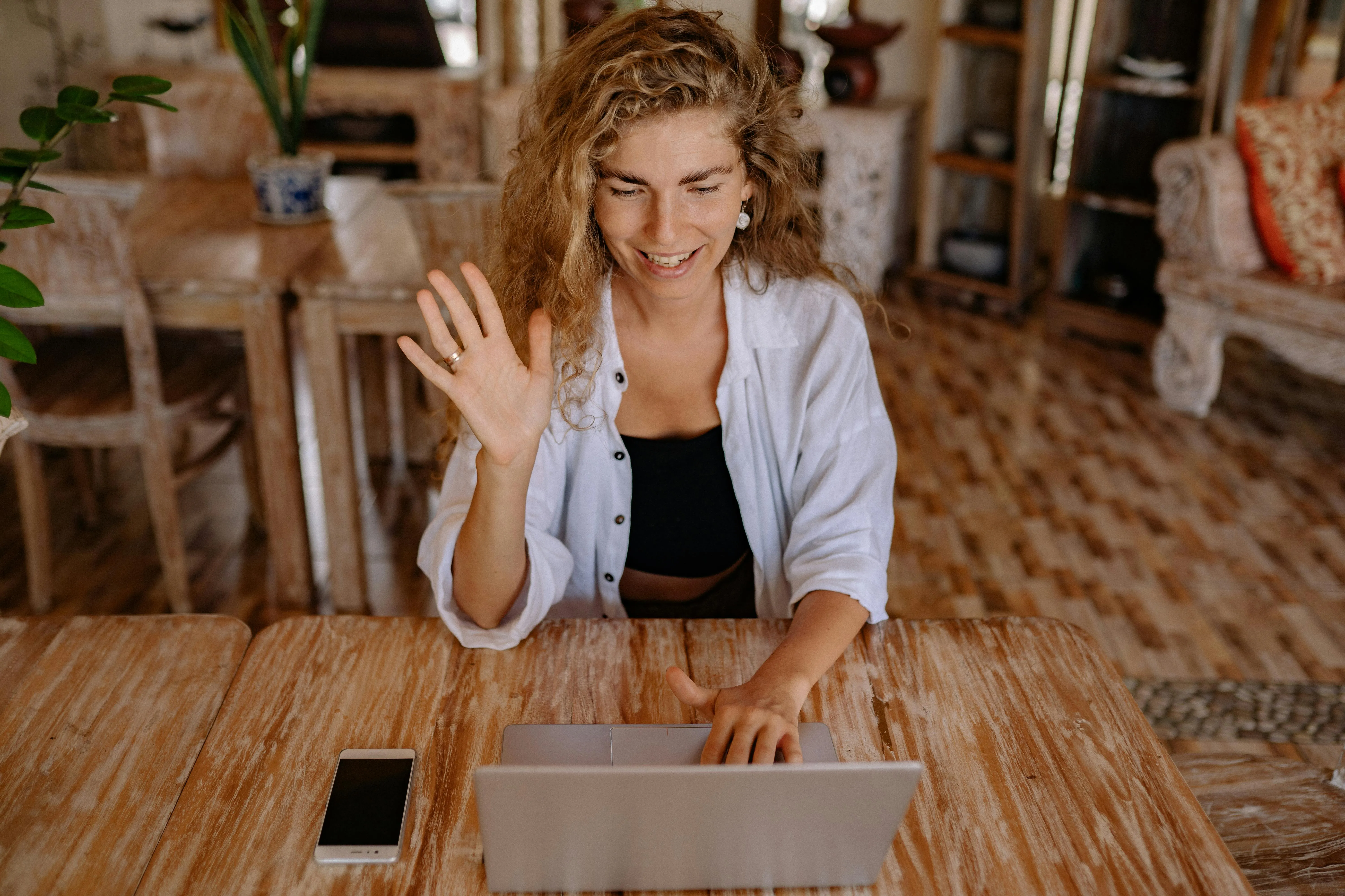 #FotoDescrição: Mulher sorridente acenando durante uma videoconferência em casa, representando interação digital e conexão.