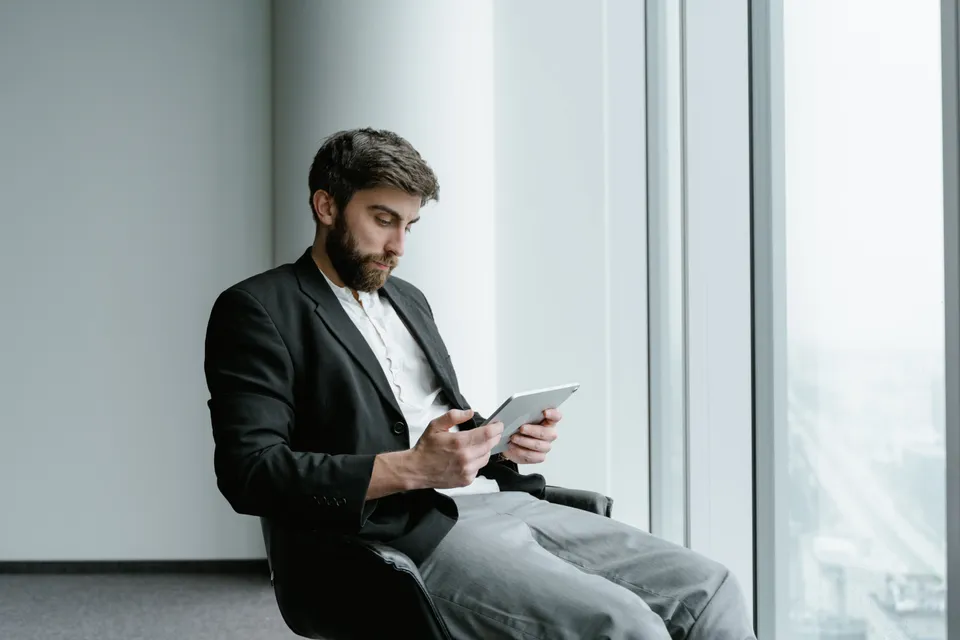 Homem sentado lendo em um tablet próximo a uma janela grande.