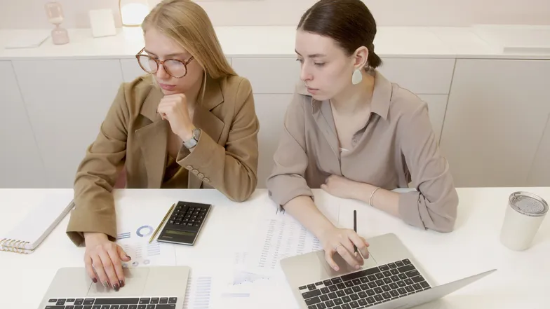 Duas mulheres analisam dados e trabalham em laptops em uma mesa.