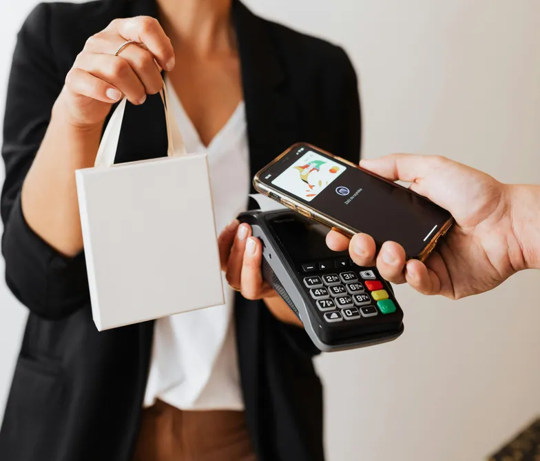 #FotoDescrição: Uma mão segurando uma sacola enquanto outra realiza pagamento digital com celular em uma máquina de cartão. Representa a integração de carteiras digitais com fidelidade.