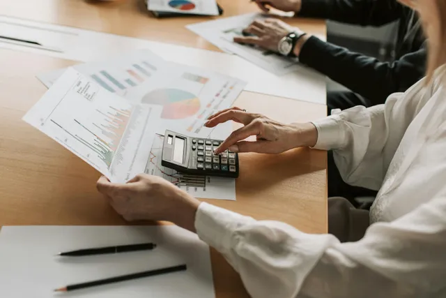 Mãos usando calculadora sobre mesa com planilhas financeiras espalhadas