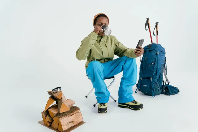 Pessoa com roupa de trilha tomando café ao lado de mochila e lenha.