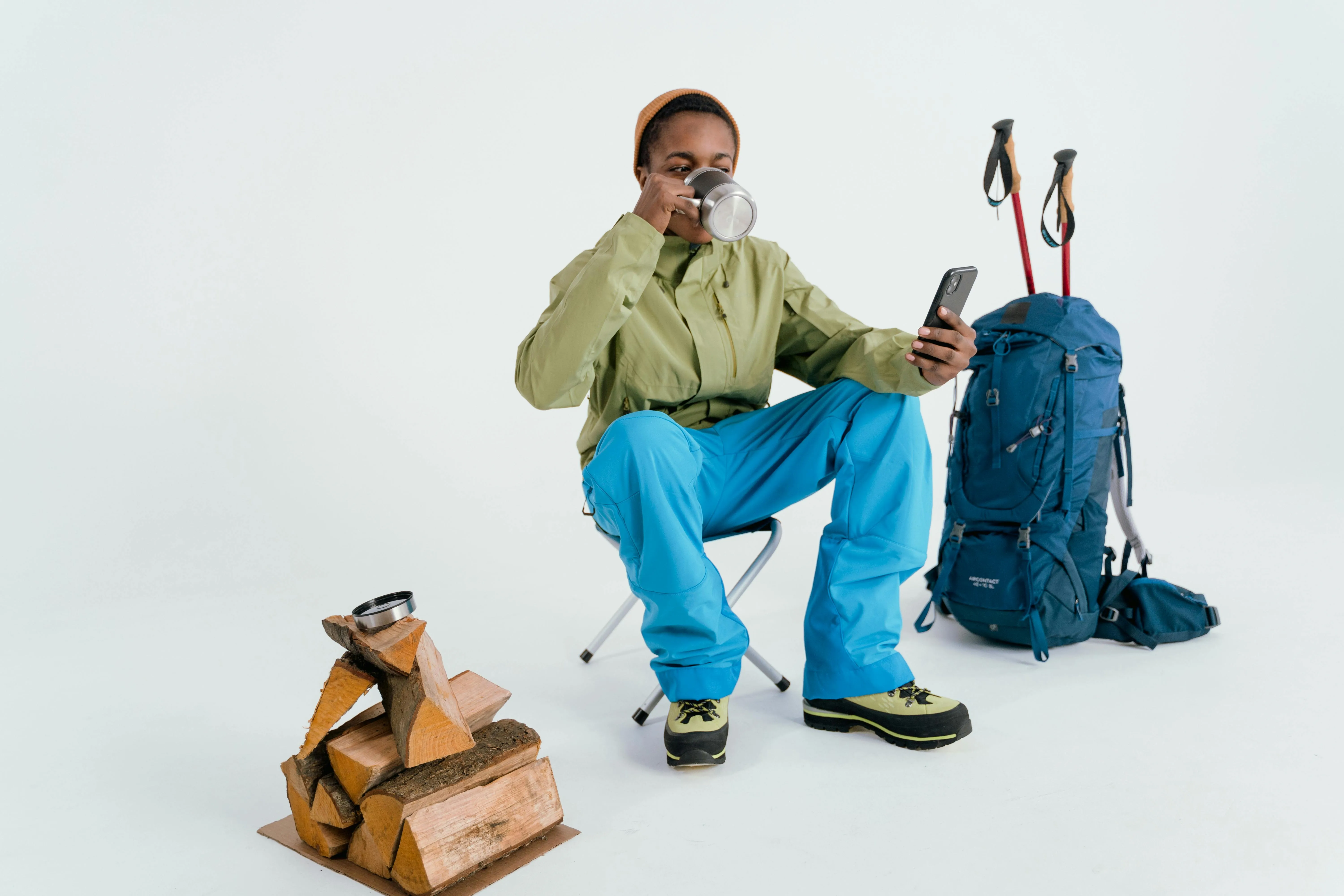 Pessoa com roupa de trilha tomando café ao lado de mochila e lenha.