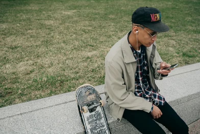 #FotoDescrição Jovem com boné e fones de ouvido sentado em um banco de pedra ao ar livre, usando o celular enquanto seu skate repousa ao lado, representando um estilo de vida urbano e conectado.