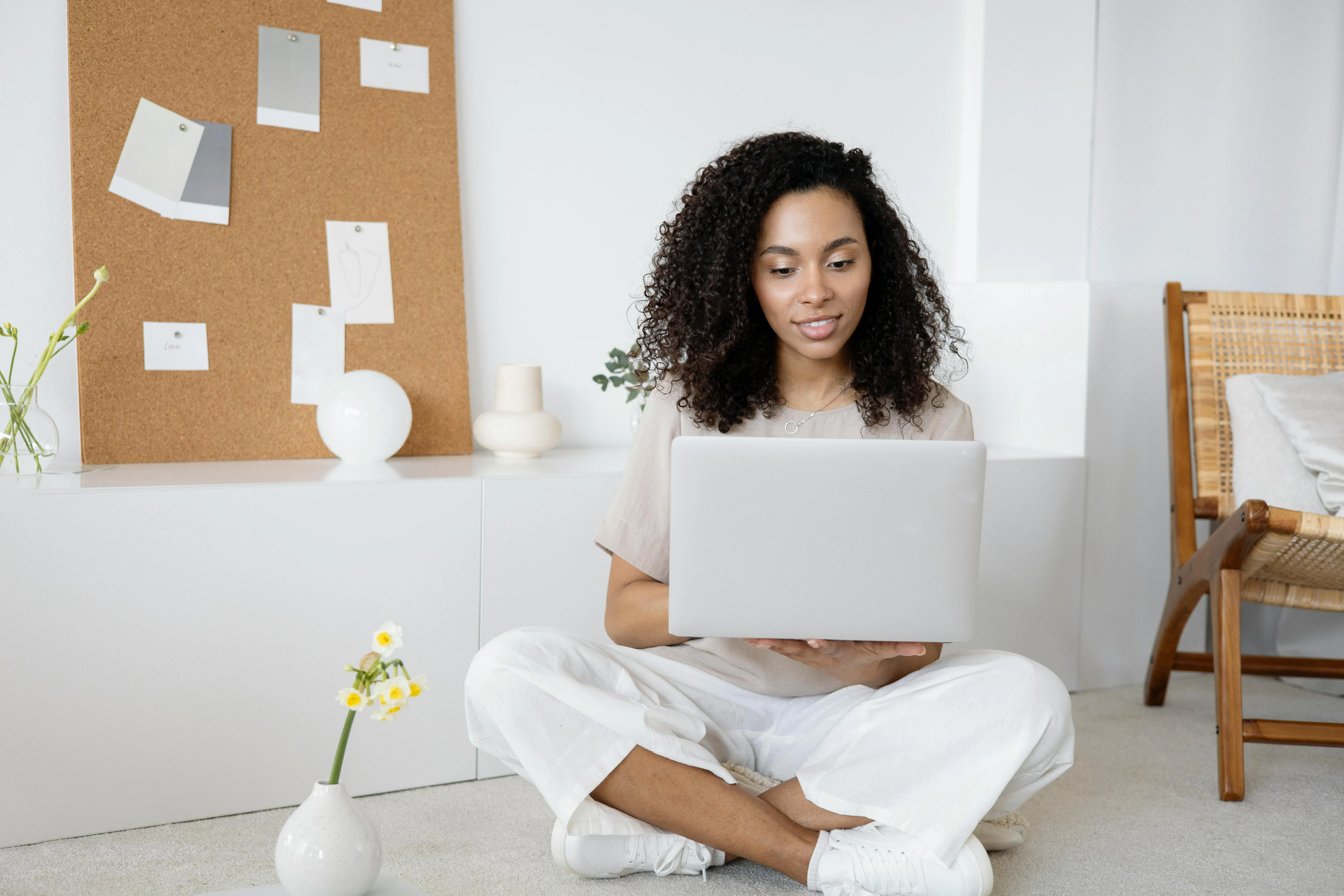 #FotoDescrição Mulher de cabelos cacheados sentada no chão com as pernas cruzadas, usando um notebook prateado em um ambiente claro com decoração minimalista.