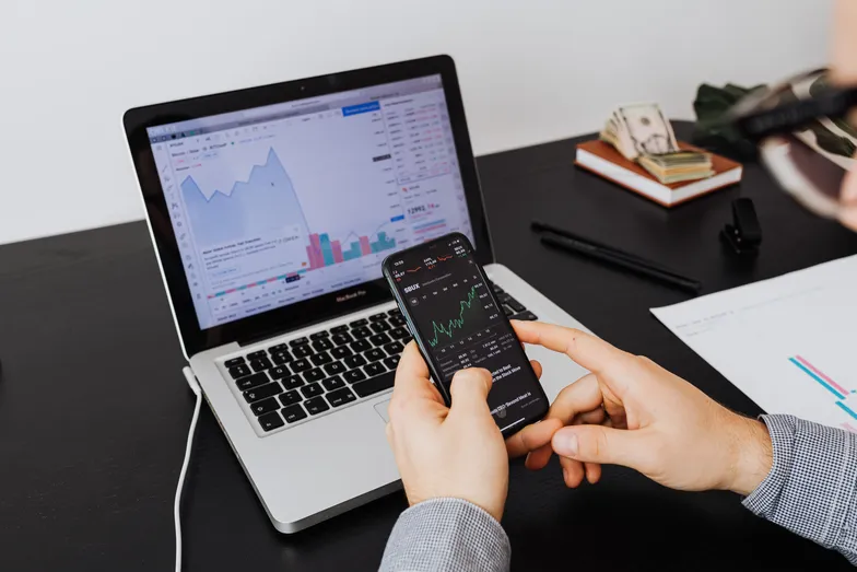 Pessoa acompanha gráficos financeiros em celular e notebook na mesa