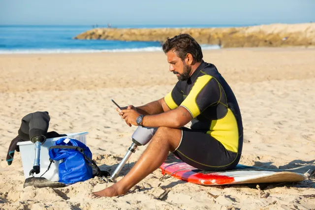 Homem com prótese na perna usa celular sentado na praia ao lado de prancha