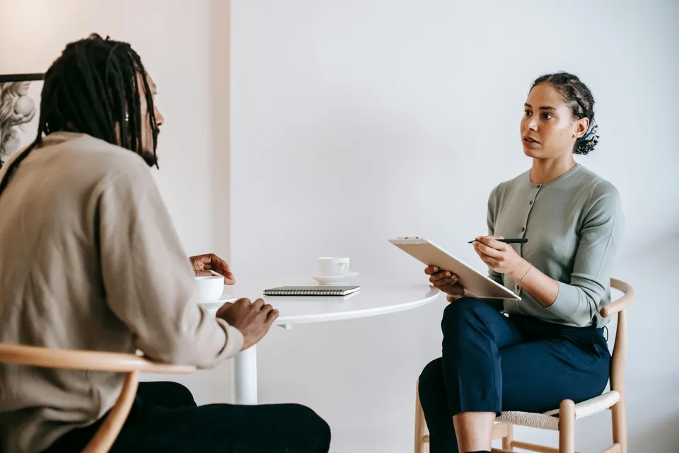 #FotoDescrição: Duas pessoas sentadas frente a frente numa mesa branca durante uma conversa, com uma mulher segurando uma prancheta e caneta, em um ambiente claro e minimalista.
