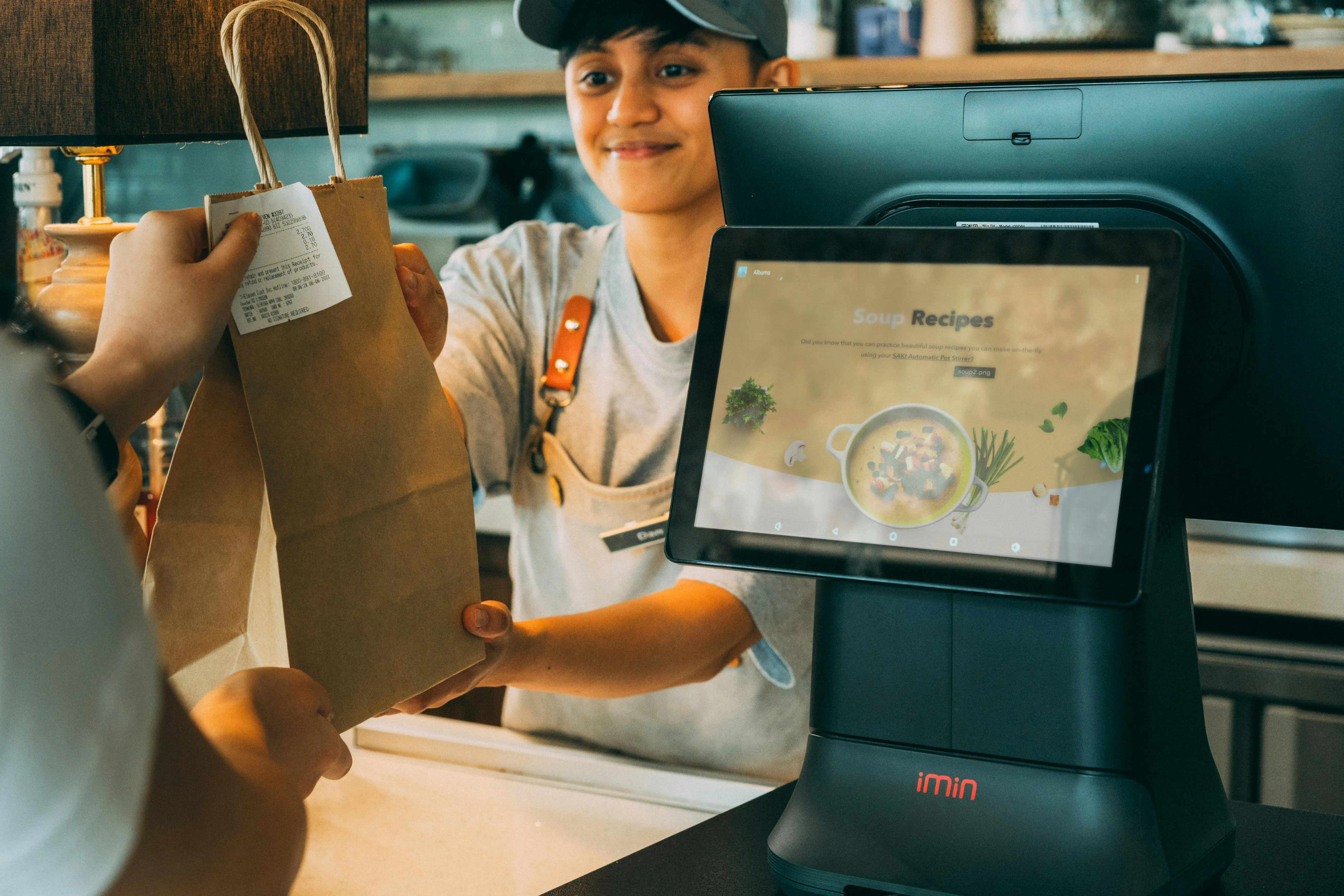 #FotoDescrição: Funcionária entregando uma sacola de papel para um cliente em um balcão de atendimento com tela de monitor.