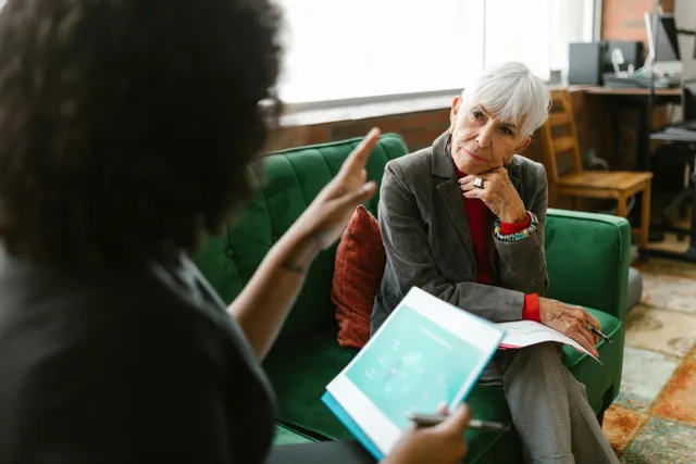 #FotoDescrição Duas mulheres conversam em um ambiente corporativo, sentadas em um sofá verde. Uma delas segura um tablet e gesticula, enquanto a outra escuta com atenção, representando uma troca estratégica sobre negócios ou relacionamento com clientes.