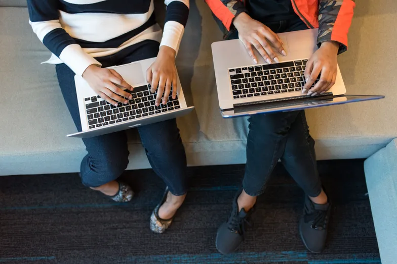 Duas pessoas usam notebooks sentadas em sofá, vista superior.