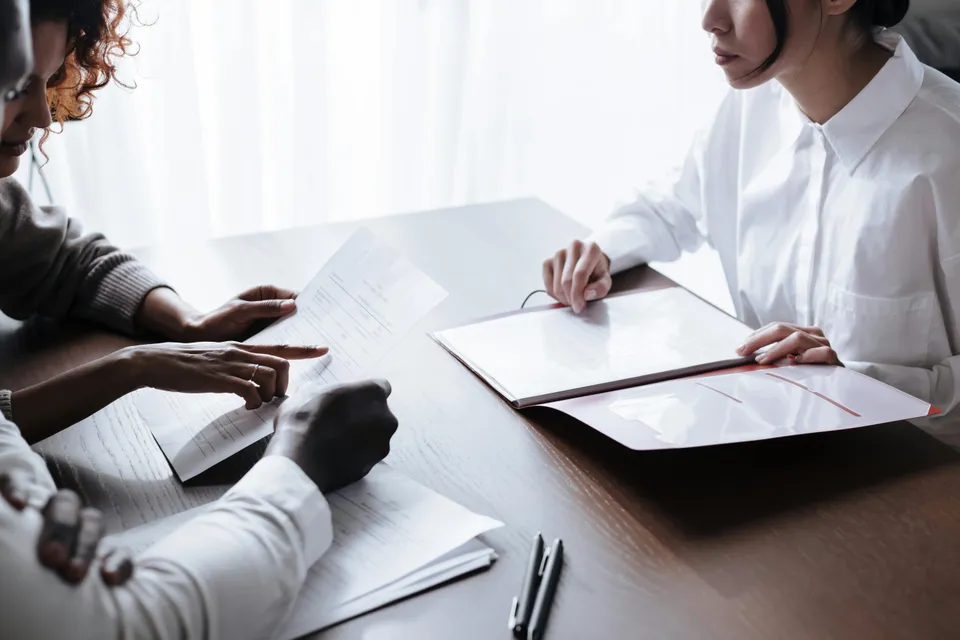 #FotoDescrição: Duas pessoas analisando documentos e contratos sobre uma mesa durante uma reunião profissional.