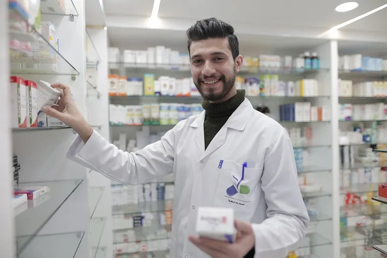 #FotoDescrição: Farmacêutico sorrindo enquanto organiza produtos em prateleiras de uma farmácia moderna e bem iluminada.