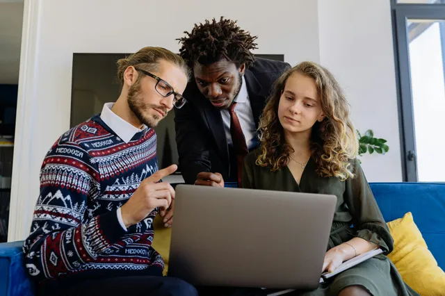 #FotoDescrição Três profissionais sentados em um sofá, observando atentamente a tela de um notebook durante uma reunião informal em um ambiente corporativo moderno.