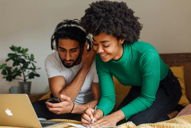 Casal estudando junto com notebook e anotações na cama.