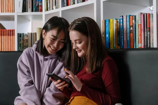 #FotoDescrição: Duas jovens sentadas em uma biblioteca olhando e sorrindo para a tela de um celular.