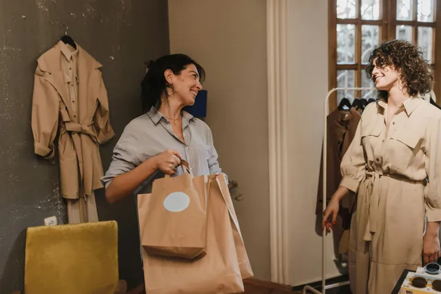 #FotoDescrição Duas mulheres sorrindo em um ambiente de loja boutique, uma delas segurando sacolas de papel, representando um momento de compra agradável e relacionamento positivo entre cliente e vendedora.